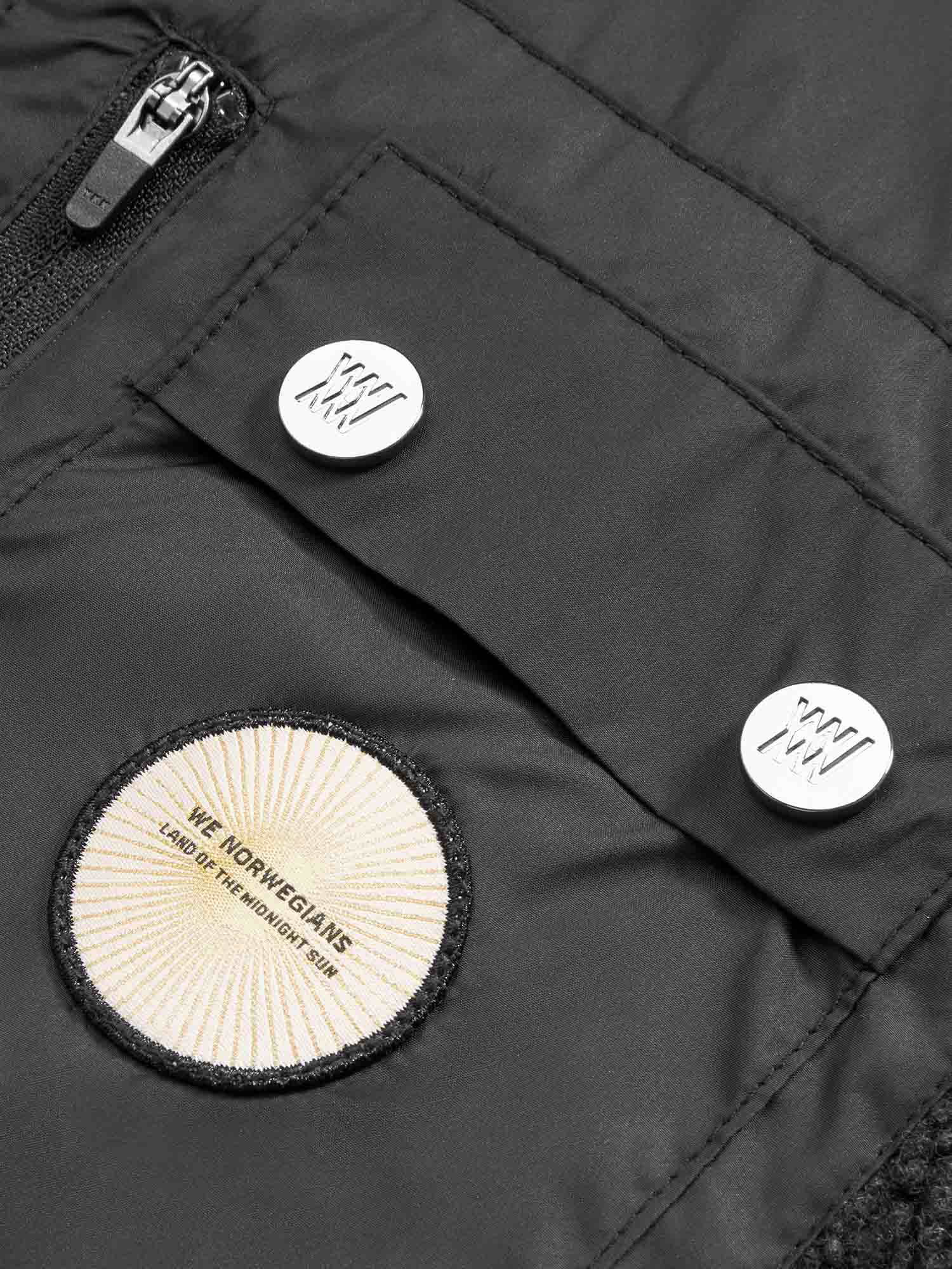 Close-up of the Alta Vest Men, a black outdoor activity vest by We Norwegians, featuring a zippered pocket, two buttons with white circular designs, and a beige patch displaying the text "WE NORWEGIANS - LAND OF THE NORTHERN RIM." The material of the jacket appears quilted.