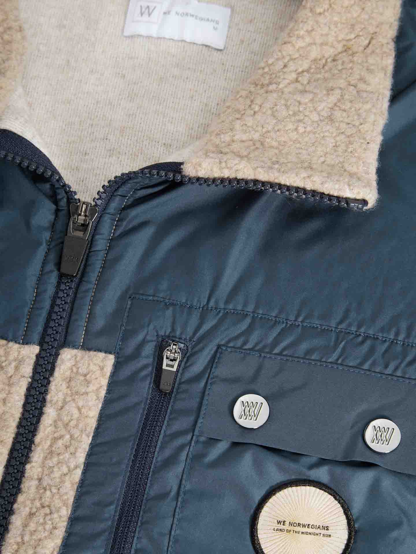 Close-up of the Alta Vest Men by We Norwegians, highlighting its blend of materials and textures. This vest features a zipper closure, a soft beige shearling collar, and dark blue fabric that evokes the look of an outdoor activity vest. Additionally, it includes a button with text and a label inside the collar area that reads "We Norwegians.