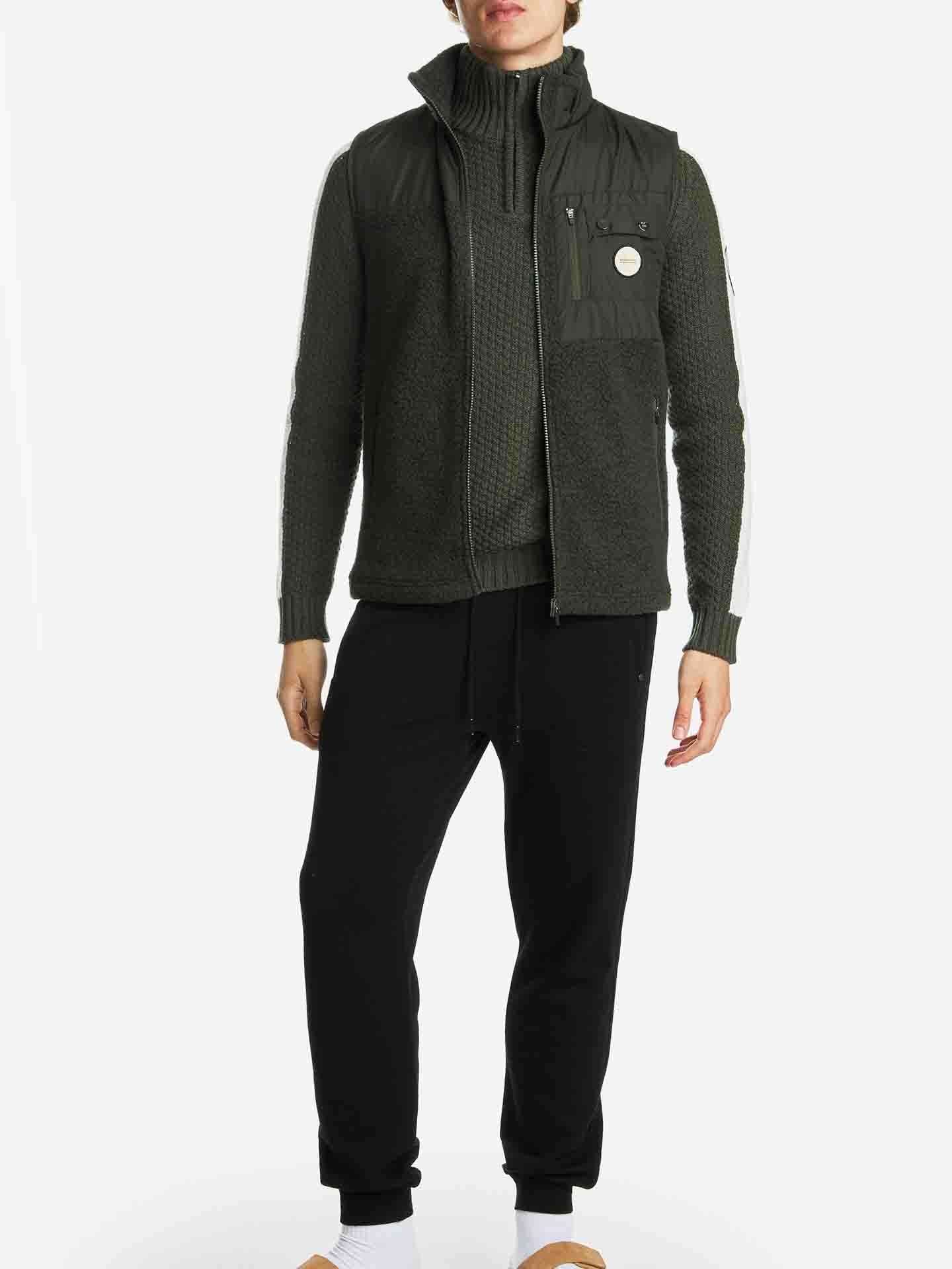 A person stands wearing a dark green quilted sweater layered with the Alta Vest Men by We Norwegians, featuring a chest pocket and a circular patch. They are also wearing black sweatpants, white socks, and brown slippers. The background is white.