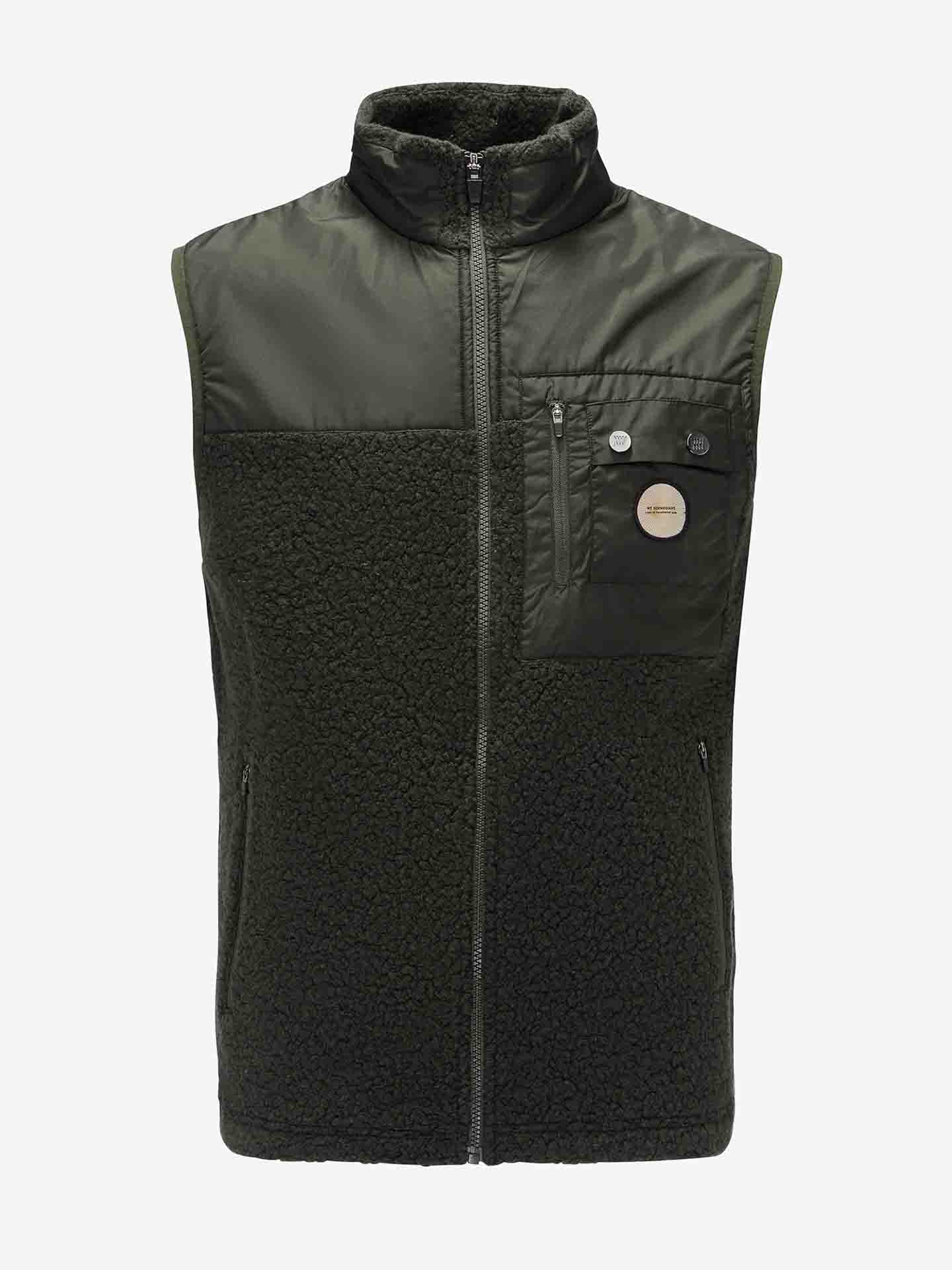 The Alta Vest Men by We Norwegians is a sleeveless, dark green vest with a zippered front and a high collar. It boasts a textured fleece-like lower half, and includes a zippered chest pocket adorned with two pins and a small logo patch. The combination of smooth and plush textures makes it the perfect après ski vest.