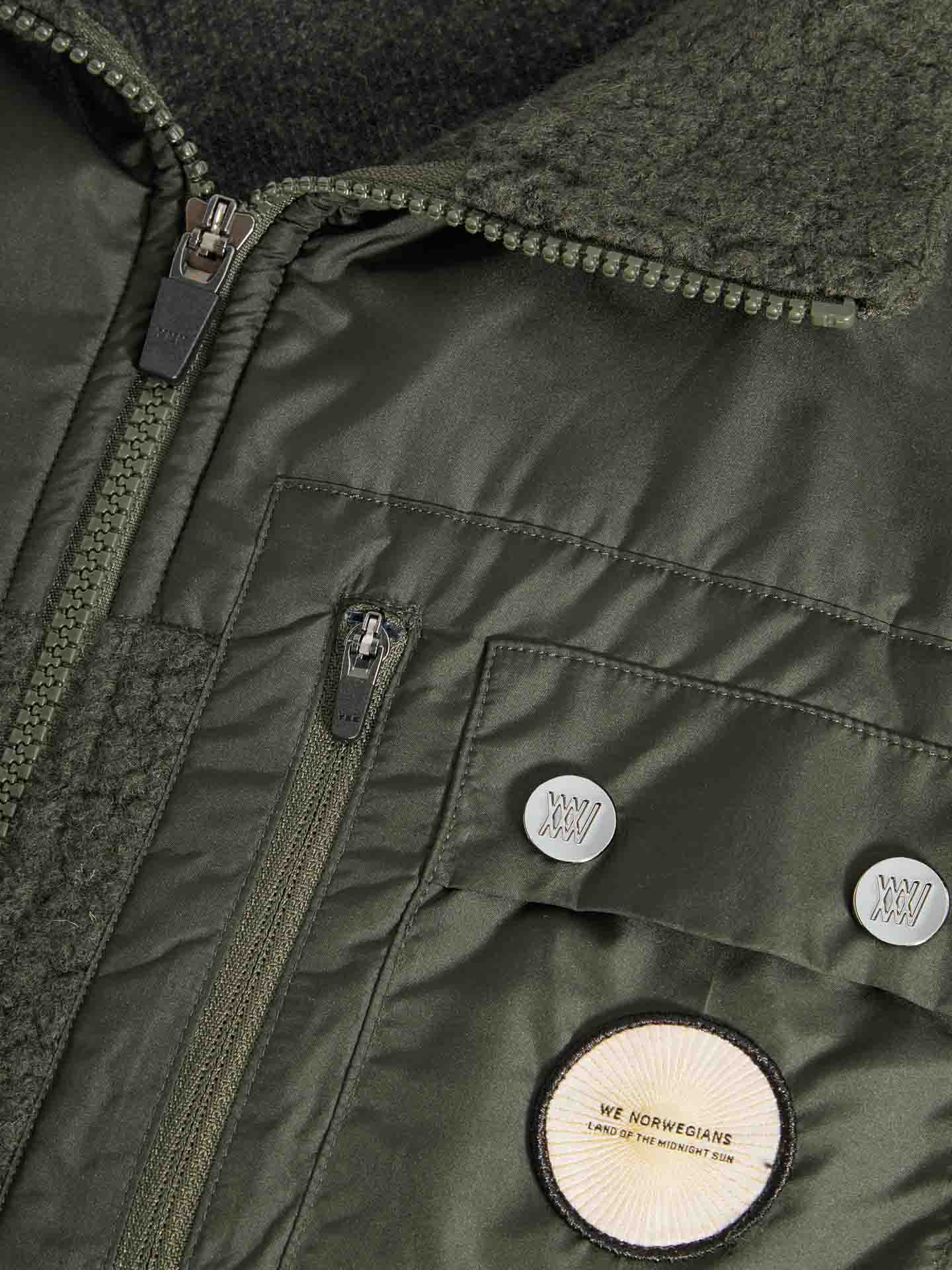 Close-up of the Alta Vest Men by We Norwegians, showcasing its green Merino wool fabric with a textured collar. The vest is designed with multiple zippers and buttons, including one that features the text "We Norwegians LAND OF THE MIDNIGHT SUN." The fabric blends smooth and fleece-like textures, making it ideal for après ski adventures.