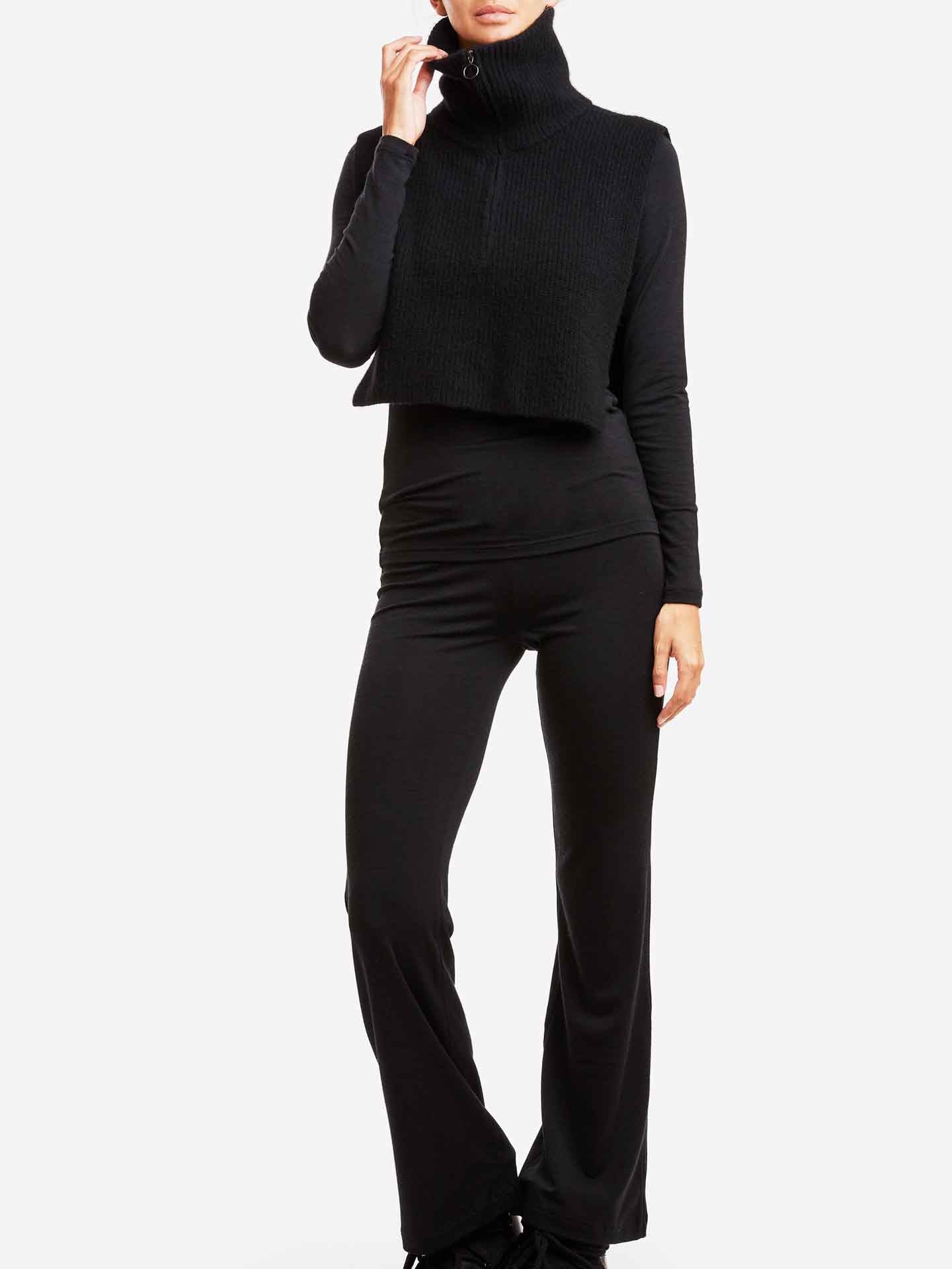 A person is standing against a plain background wearing a black, long-sleeve top, Hygge Flared Pant Women by We Norwegians, and a cropped sleeveless black turtleneck sweater. The person's left hand is raised, touching the collar of the HYGGE base layer turtleneck. Their face is partially out of frame.