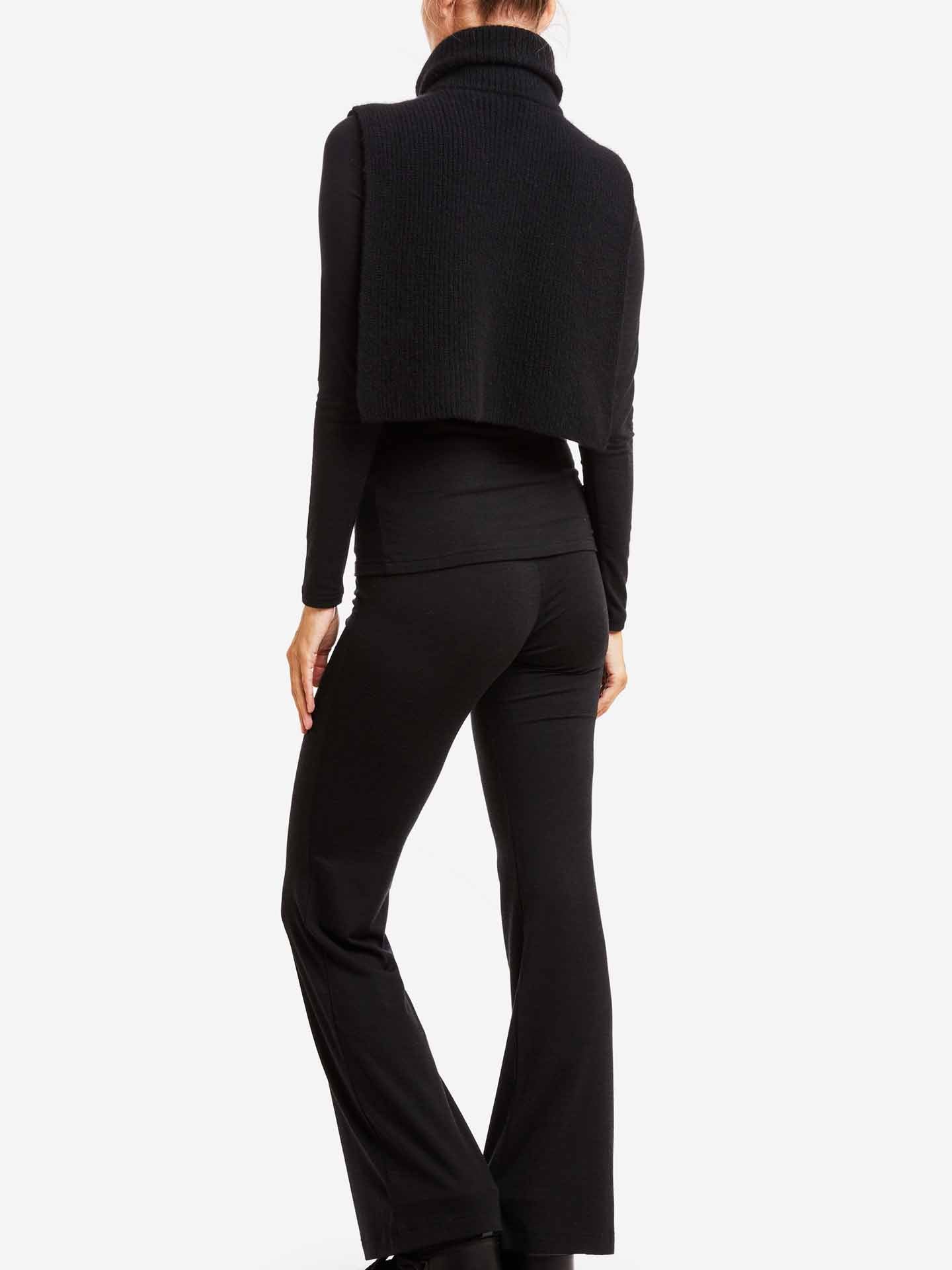 A person is seen from the back, wearing a black turtleneck sweater, Hygge Flared Pant Women by We Norwegians, and a sleeveless knit vest made of merino wool. The form-fitting outfit showcases a relaxed stance against a white background.