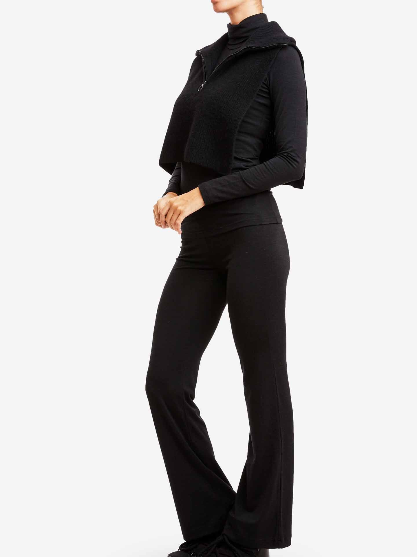 A person is standing sideways, wearing a black long-sleeve merino wool top with a zip-up vest draped over their shoulders and We Norwegians' Hygge Flared Pant Women in black. The background is plain white, highlighting the outfit.