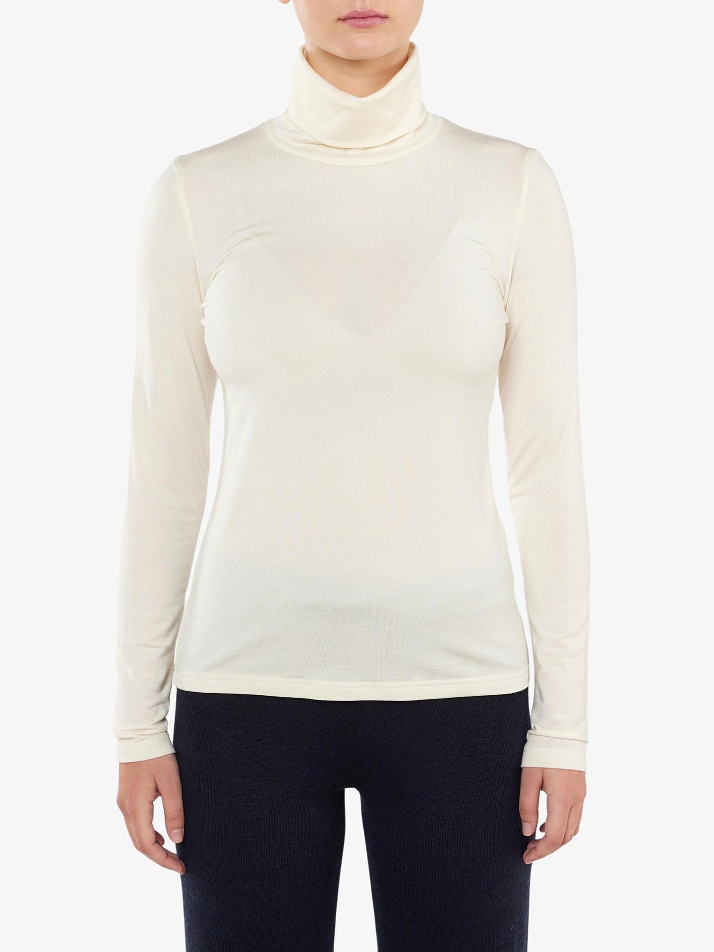 A person wearing a long-sleeved Hygge Turtleneck Women from We Norwegians, an off-white top made of merino wool, paired with black pants stands against a plain white background. Only the upper body and arms are visible, with the head not fully shown.