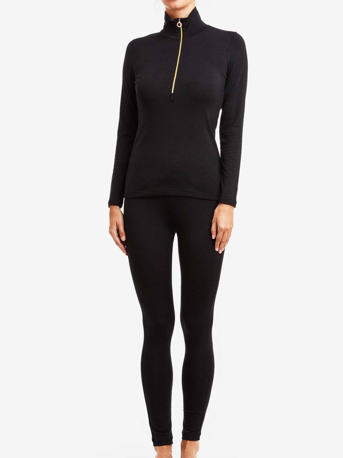 A person is wearing the Hygge Zip Up Women by We Norwegians, a fitted black outfit consisting of a long-sleeve top with a small front zipper and matching leggings. The luxury layering piece stands out against the plain white background, with their arms relaxed at their sides. The outfit appears to be made of stretchy material.