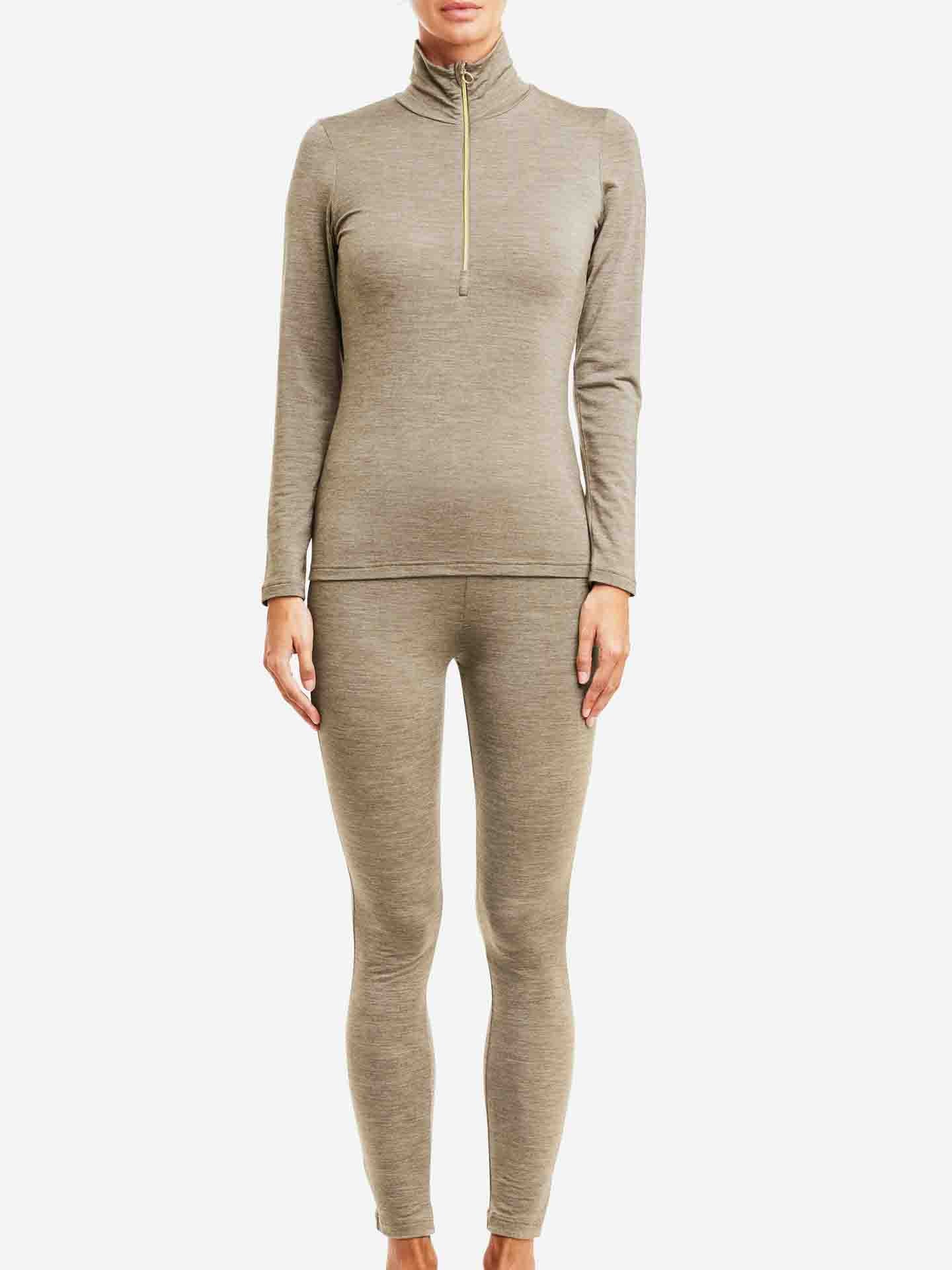 A person wearing the We Norwegians Hygge Zip Up Women, a form-fitting, long-sleeve zipper top in light brown with matching leggings, stands barefoot against a plain white background. The outfit appears to be made of soft, stretchy merino wool fabric suitable for active wear or lounging, serving as both a luxury layering piece and Hygge base layer.