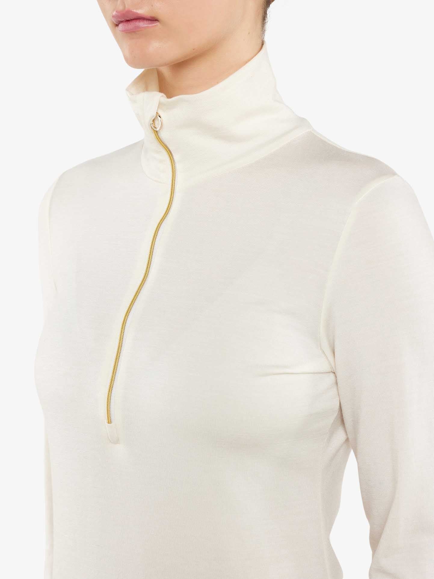 Close-up of a person wearing the Hygge Zip Up Women by We Norwegians. The off-white turtleneck sweater features a gold zipper running from the collar to mid-chest. The fabric, a blend of Tencel and Merino wool, appears soft and slightly sheer. The head is partially visible, emphasizing the luxury base layer's upper body and garment details.