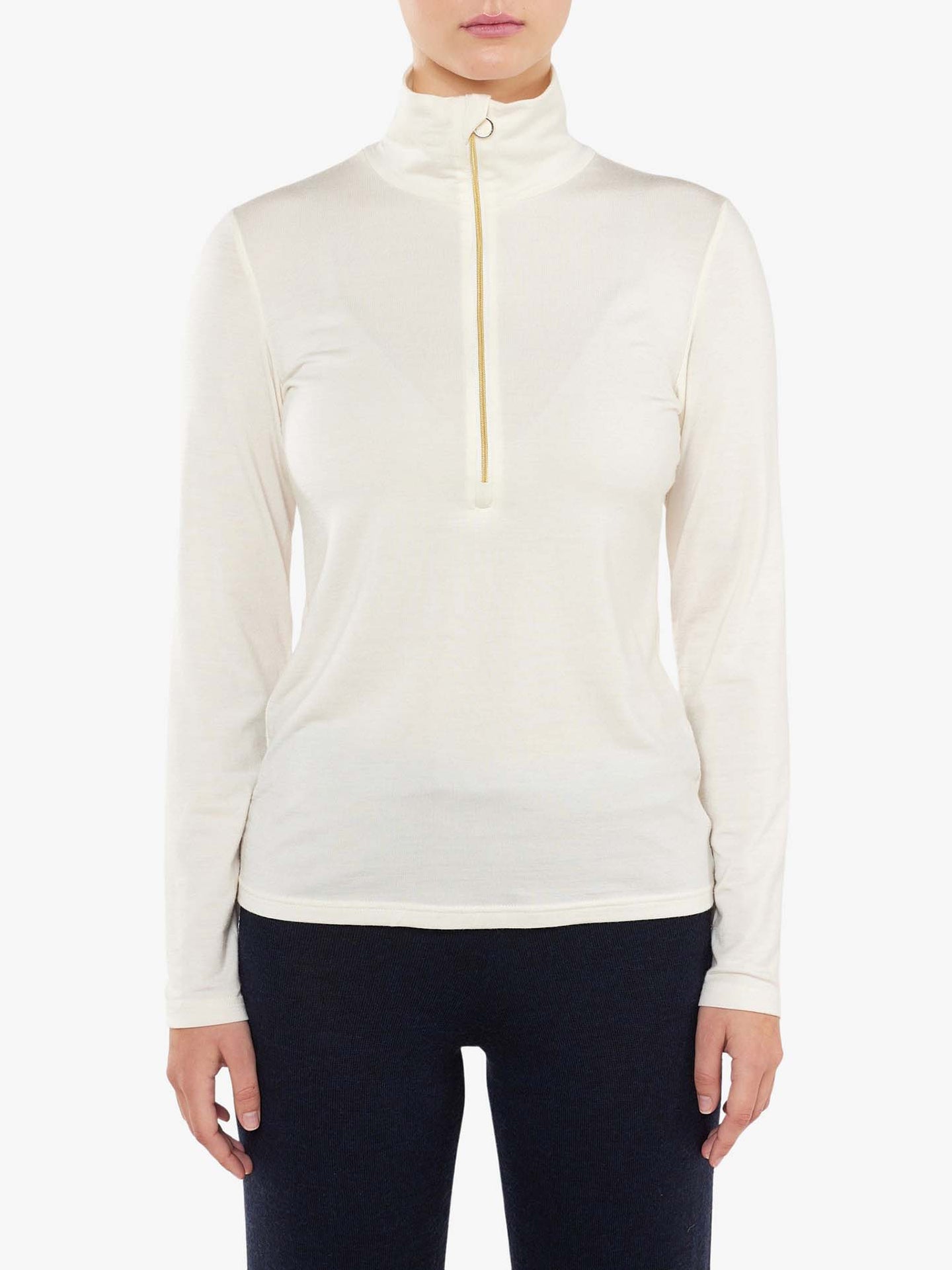 A person, dressed in the We Norwegians Hygge Zip Up Women base layer with long sleeves and an off-white hue, stands against a plain white background. The Merino wool top features a high collar and a zipper extending to the chest. The individual pairs it with black pants in a photo cropped from the neck down to the hips.