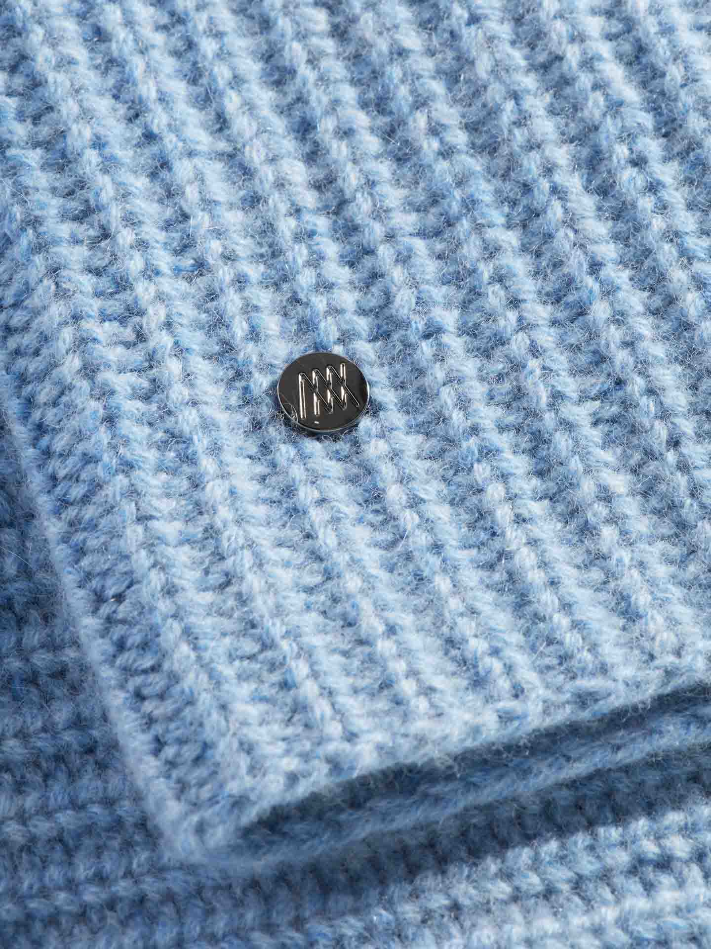 A close-up view of the light blue, ribbed knit fabric of the Blefjell Cashmere Neckwarmer by We Norwegians reveals a metallic button with a small geometric design. The texture and cashmere feel are clearly visible, showcasing its soft and cozy appearance.