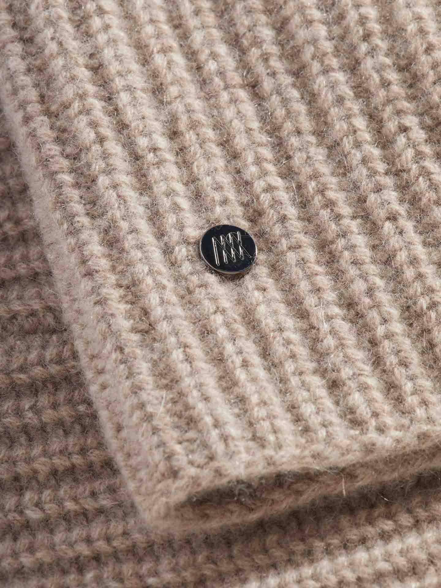 Close-up of a beige knitted fabric with a ribbed texture, reminiscent of luxurious Blefjell Cashmere Neckwarmers from We Norwegians. A small, round, silver button with an engraved pattern is sewn onto the fabric, adding a subtle detail to the knit.