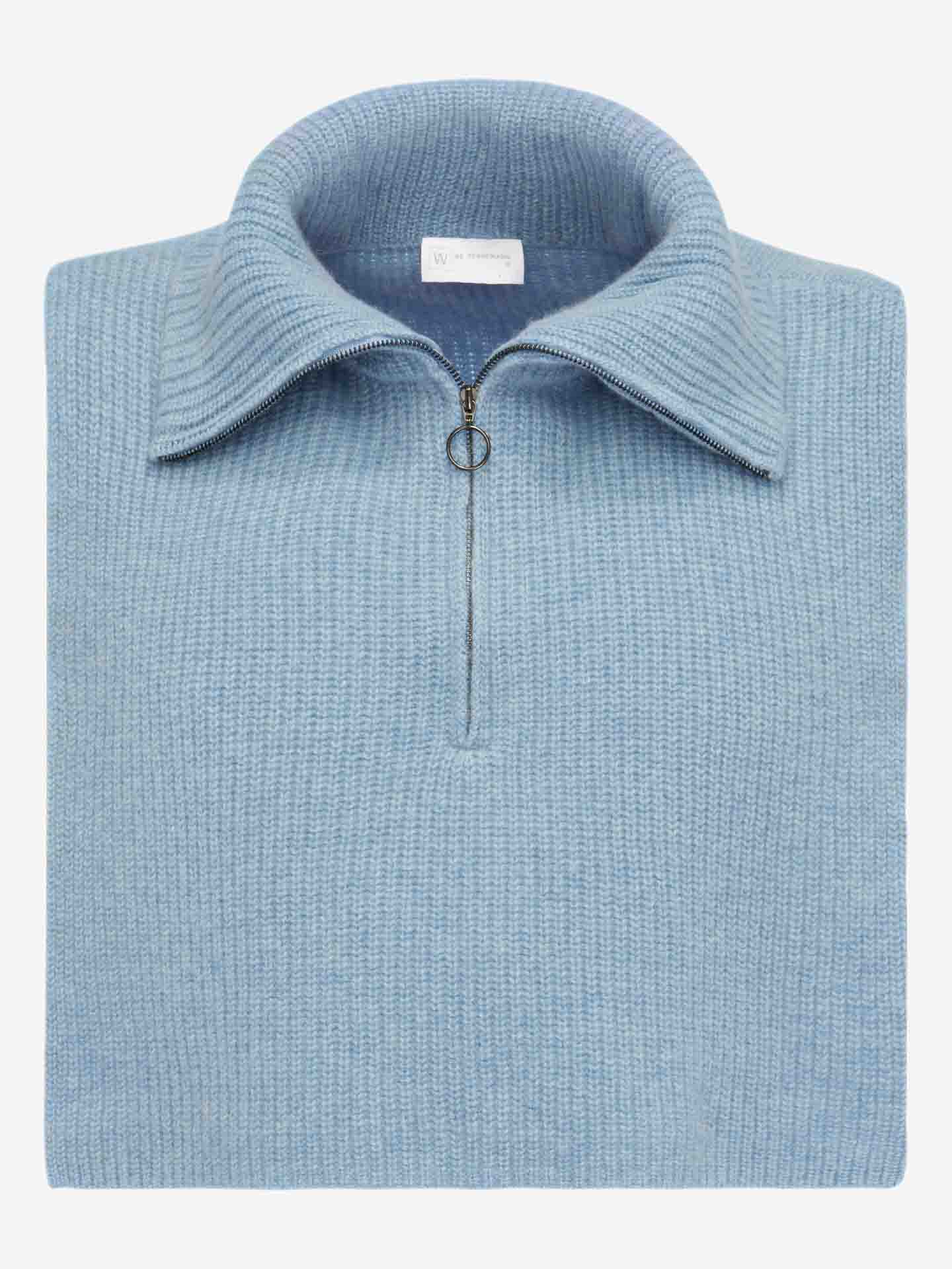 A luxuriously folded knit sweater in light blue, featuring a cashmere bib and a metal half-zip with a circular zipper pull, is displayed against a plain white background. The collar is neatly turned up, showcasing the label of We Norwegians' Blefjell Bib Women at the top inside the collar.