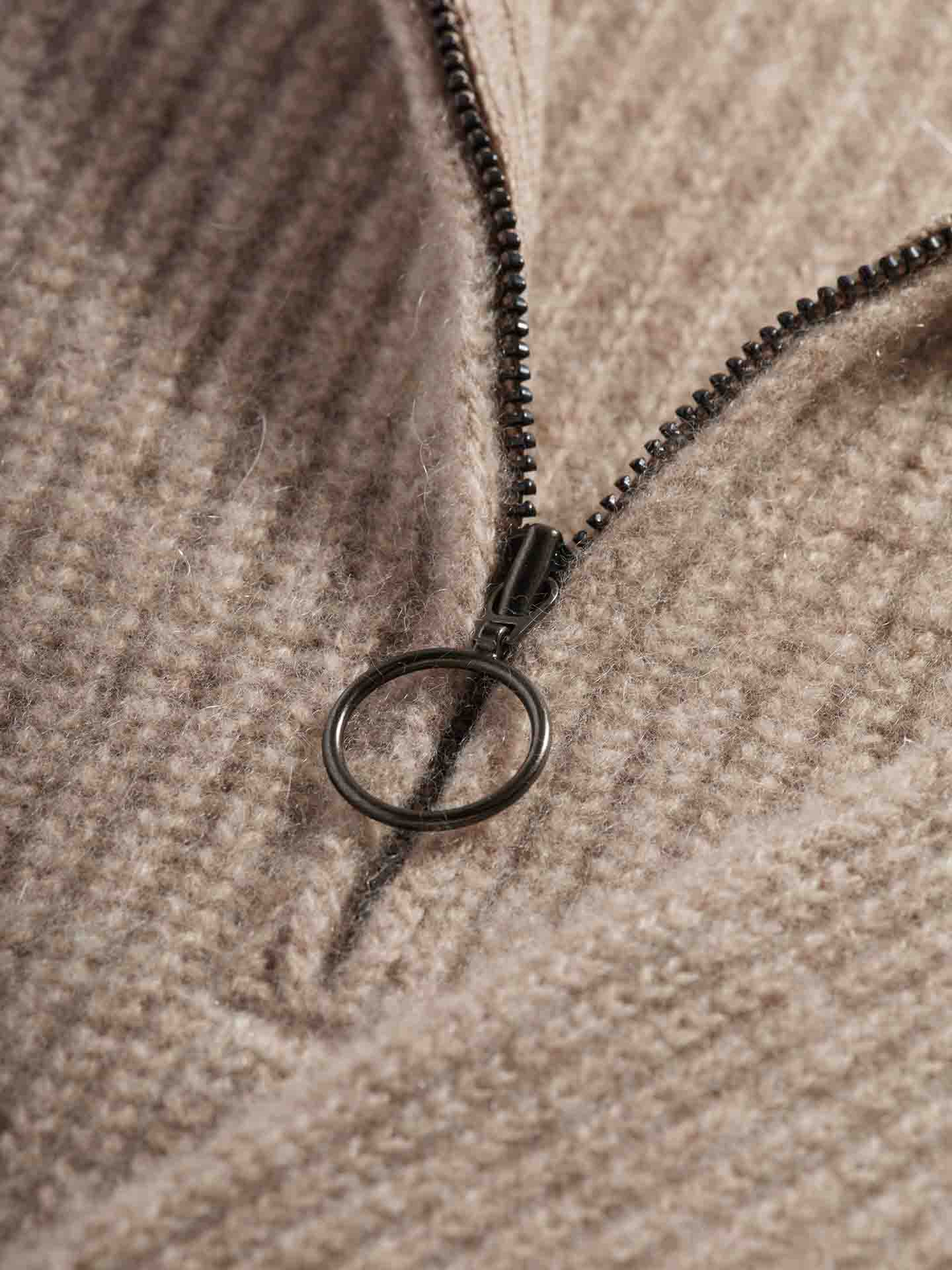 Close-up of the textured beige Blefjell Bib Women by We Norwegians, featuring a zippered closure. The zipper is adorned with a round metal pull tab, which elegantly contrasts with the ribbed knit fabric, creating a sophisticated designer vest aesthetic.