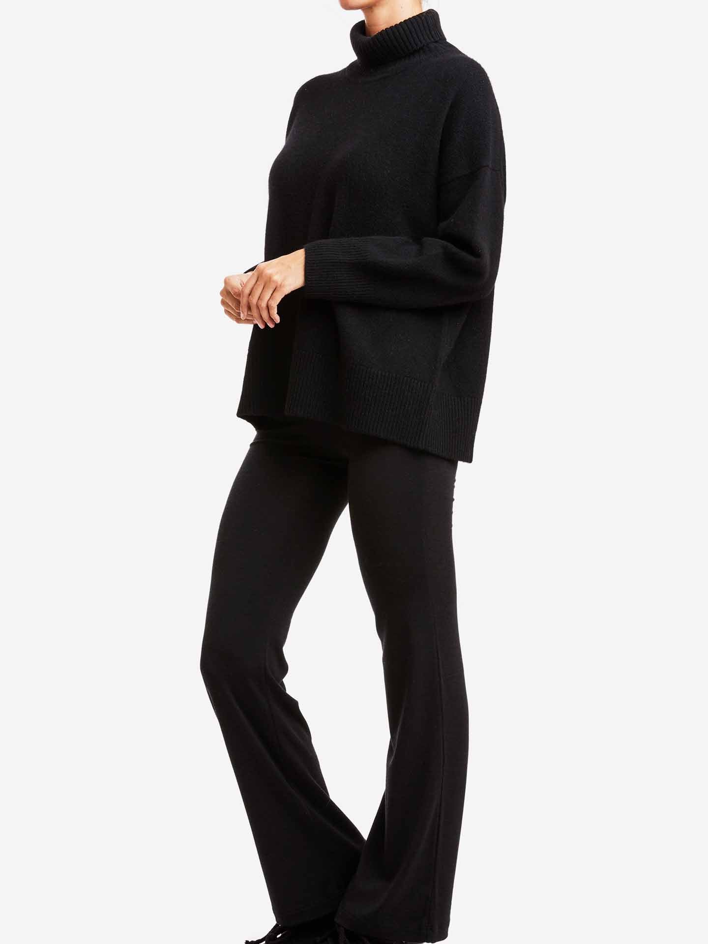 A person is standing against a plain white background, dressed in We Norwegians' Blefjell Sweater Women—a black, long-sleeved, high-neck cashmere sweater paired with black pants. The outfit appears comfortable and boasts an oversized fit. The person's head is not visible in the image.