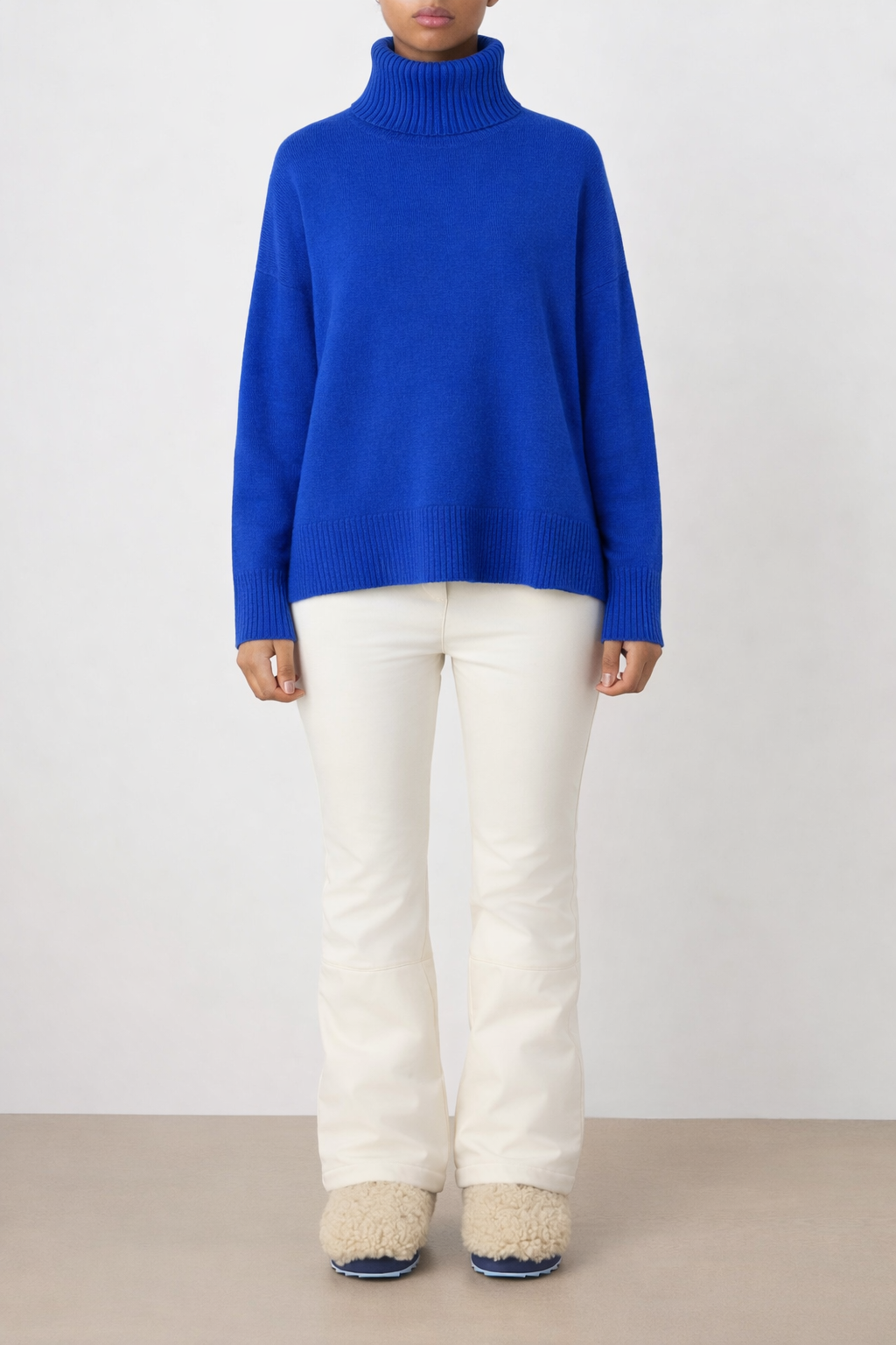 A person stands against a light background wearing the We Norwegians Blefjell Sweater Women in bright blue cashmere, paired with off-white pants and fluffy off-white shoes. Their arms are relaxed by their sides; face is not visible.