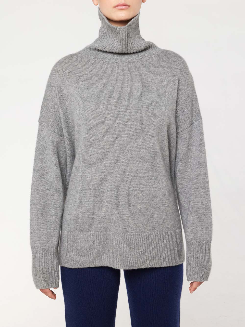 A person is wearing the We Norwegians Blefjell Sweater Women, a gray turtleneck with long sleeves that extend over the wrists, paired with dark-colored pants. The background is plain and light-colored, accentuating the cozy, oversized fit of the cashmere sweater.