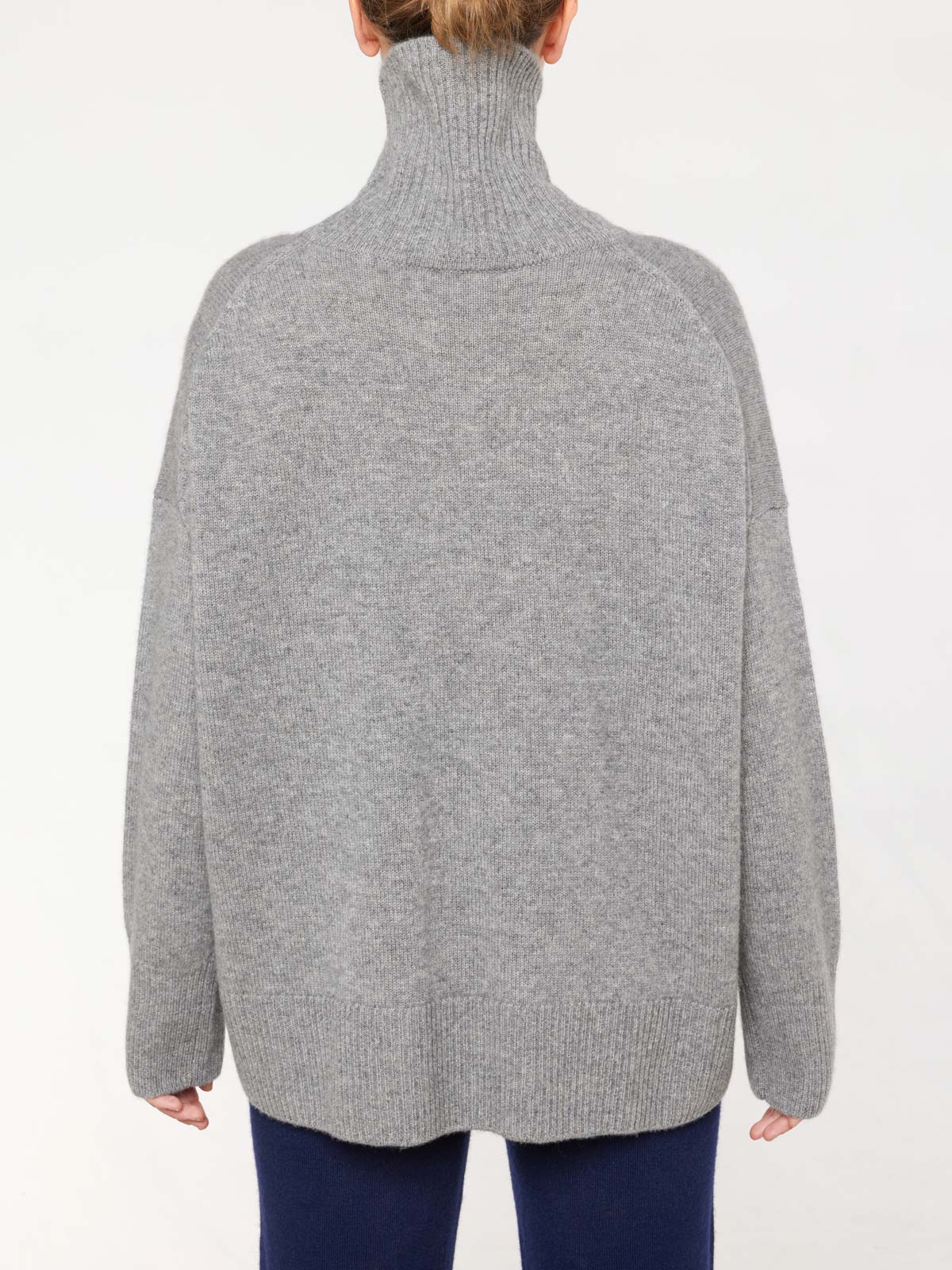 A person clad in the We Norwegians Blefjell Sweater Women, an oversized gray cashmere piece with a loose-fitting turtleneck and ribbed hem and cuffs, is photographed with their back facing the camera. Paired with dark blue pants, the luxurious ensemble stands out against a plain white background.