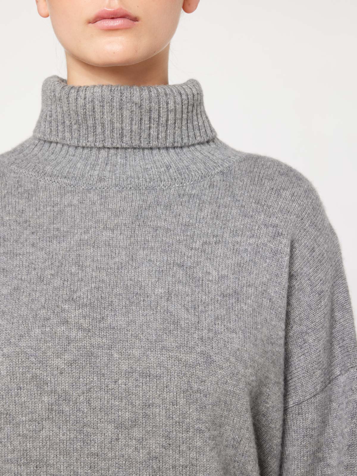 A person wearing the luxurious We Norwegians Blefjell Sweater Women in grey. The photo is cropped to show only the lower part of their face and upper torso, highlighting the oversized fit with its thick, ribbed collar and soft, cozy texture. The background is plain and light-colored.