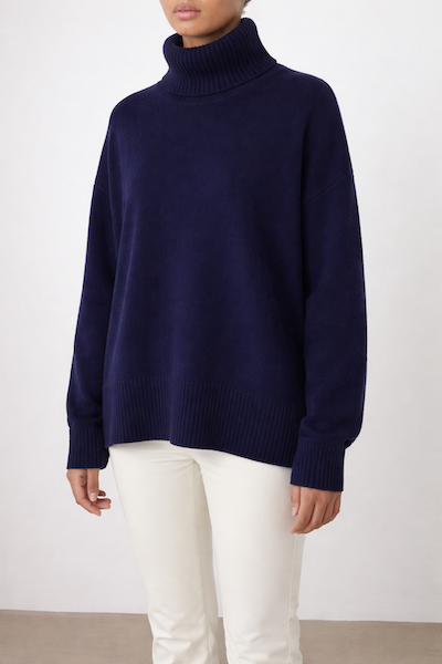 A woman wears the We Norwegians Blefjell Sweater Women - Pure Cashmere, a loose-fitting navy blue turtleneck with ribbed cuffs and hem, paired with white pants against a neutral background.