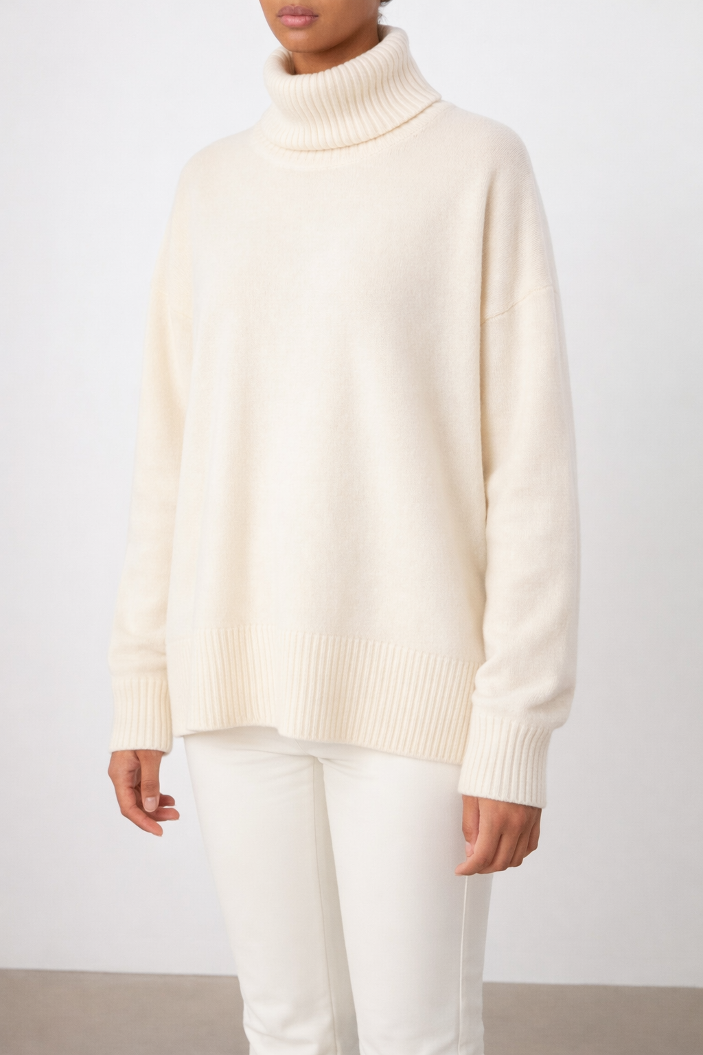 A person wears the We Norwegians Blefjell Sweater Women—Pure Cashmere in ivory, featuring a relaxed fit with ribbed cuffs, hem, and neck. She stands against a plain light background, her face partially out of frame.