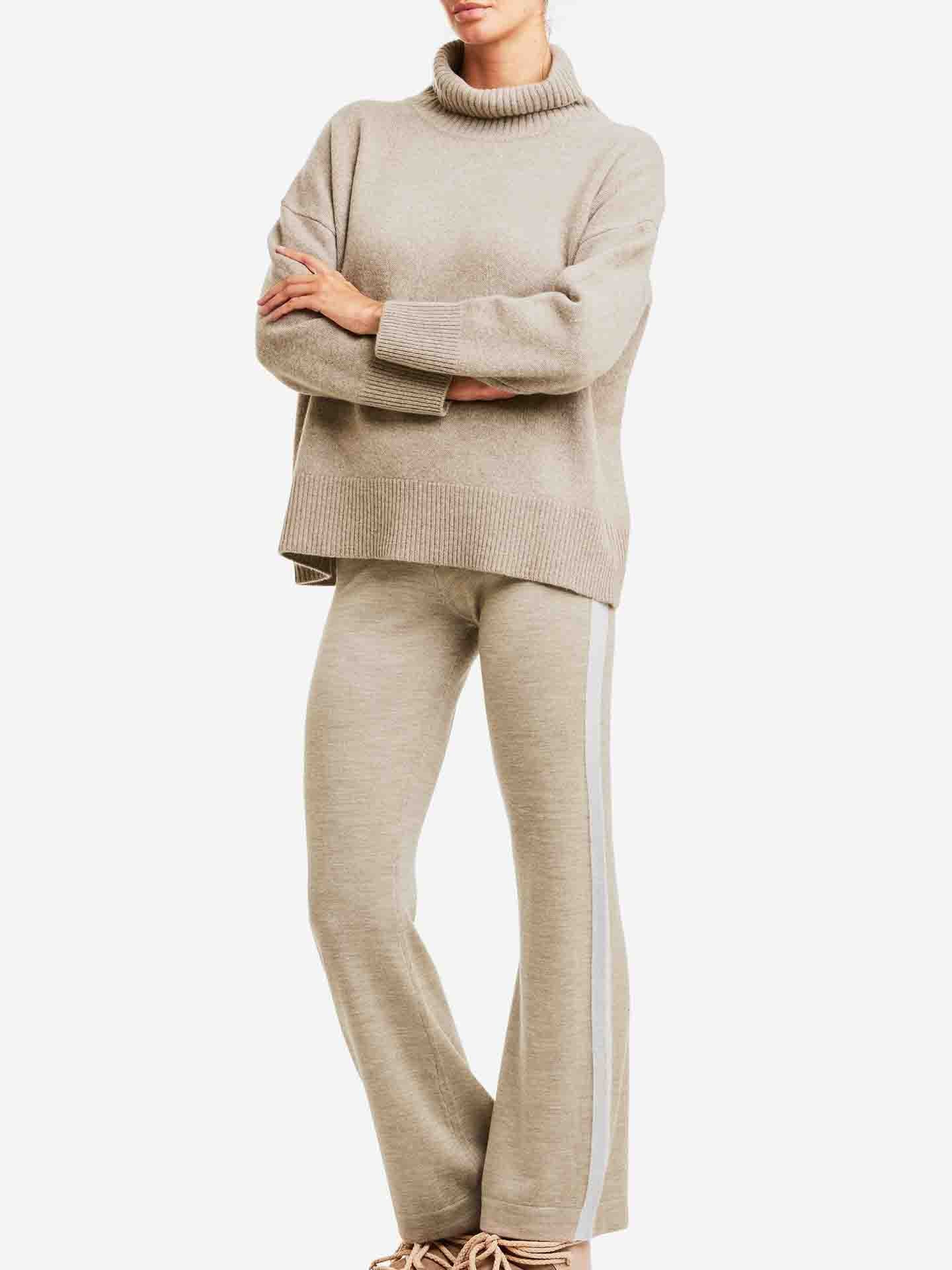 A person stands with arms crossed, wearing a cozy, oversized Blefjell Sweater Women by We Norwegians and matching beige pants with white side stripes. They have casual, light-colored sneakers and are against a white background, exuding a sense of affordable luxury.