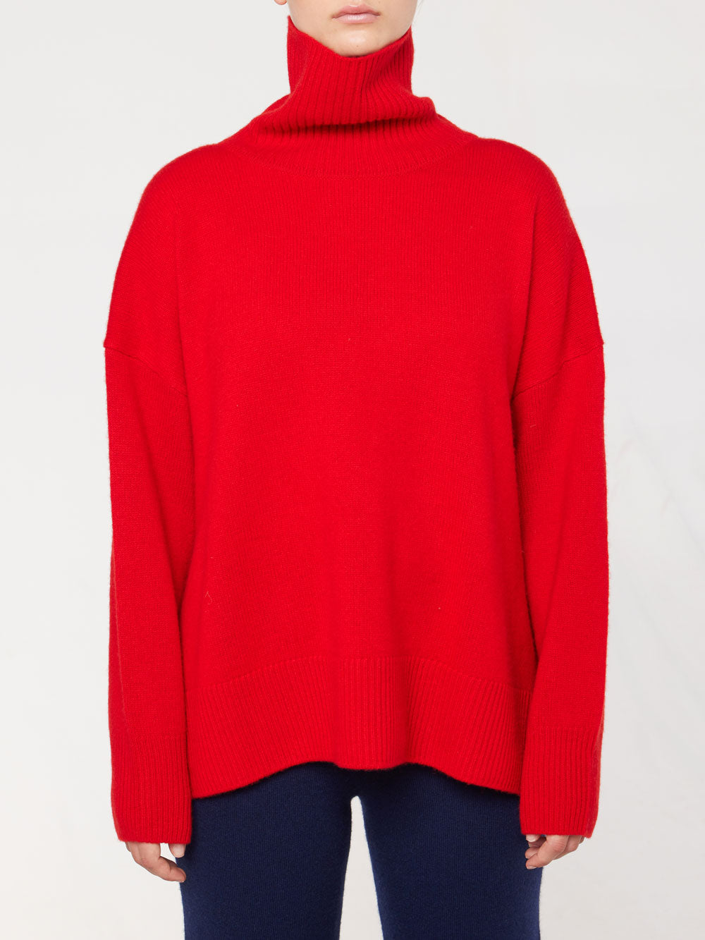 A person wearing the Blefjell Sweater Women by We Norwegians—a bright red, cashmere turtleneck sweater with long sleeves. The luxurious piece features an oversized fit and extends past the waist. The individual is also wearing dark pants against a plain, light-colored background.