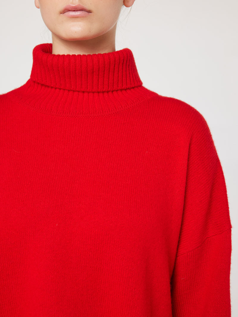 A person is wearing the We Norwegians Blefjell Sweater for women, a bright red, chunky knit turtleneck with an oversized fit. The focus is on the sweater and the lower part of the person's face, set against a plain, light grey background.