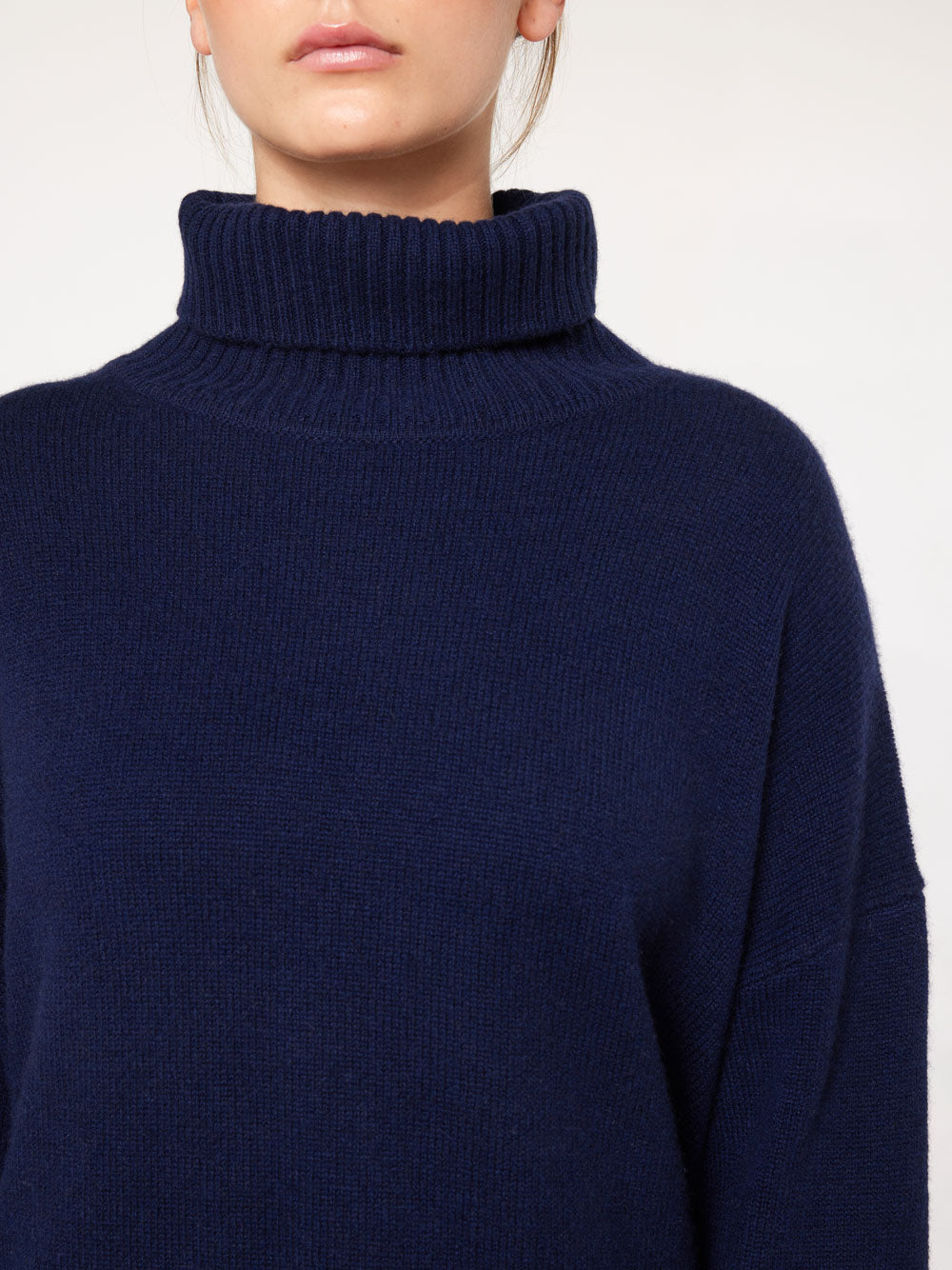 A person wearing a Blefjell Sweater Women by We Norwegians—it's a navy blue, long-sleeved, cashmere turtleneck sweater. The image focuses on the upper torso and neck, cutting off part of the person's face and showing only the lower half of their lips and chin. The background is a plain off-white color.