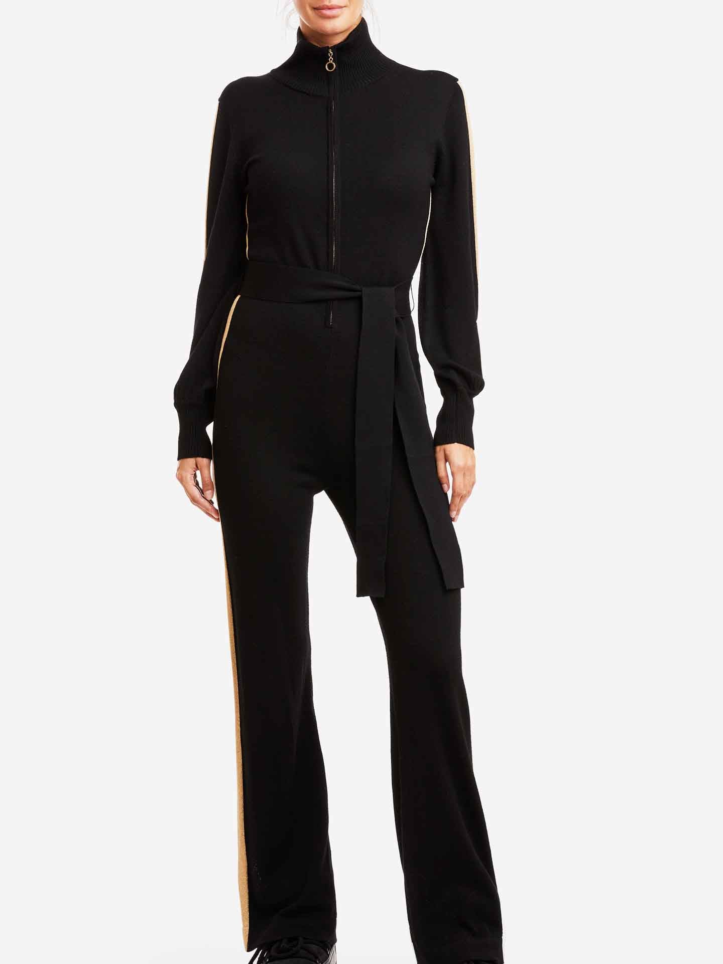 A person wearing the chic Geilo Jumpsuit Women by We Norwegians, made of luxurious Merino Wool, featuring a zippered front and a waistband tie. The black, long-sleeved jumpsuit boasts light-colored stripes on the sides of the arms and legs. The individual is standing against a plain white background, exuding elegance.