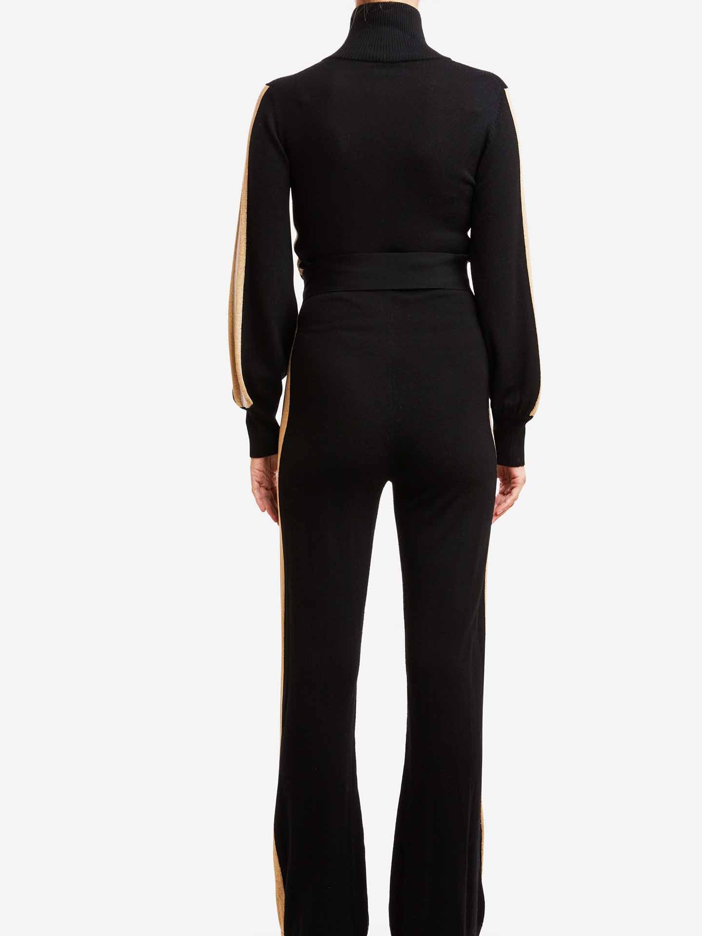 A person is standing with their back to the camera, donning the stylish Geilo Jumpsuit Women by We Norwegians. This black outfit, crafted from luxurious merino wool, features long sleeves, a high neck, and wide-legged pants, all enhanced by gold stripes running along the sides. The backdrop is a simple white color.