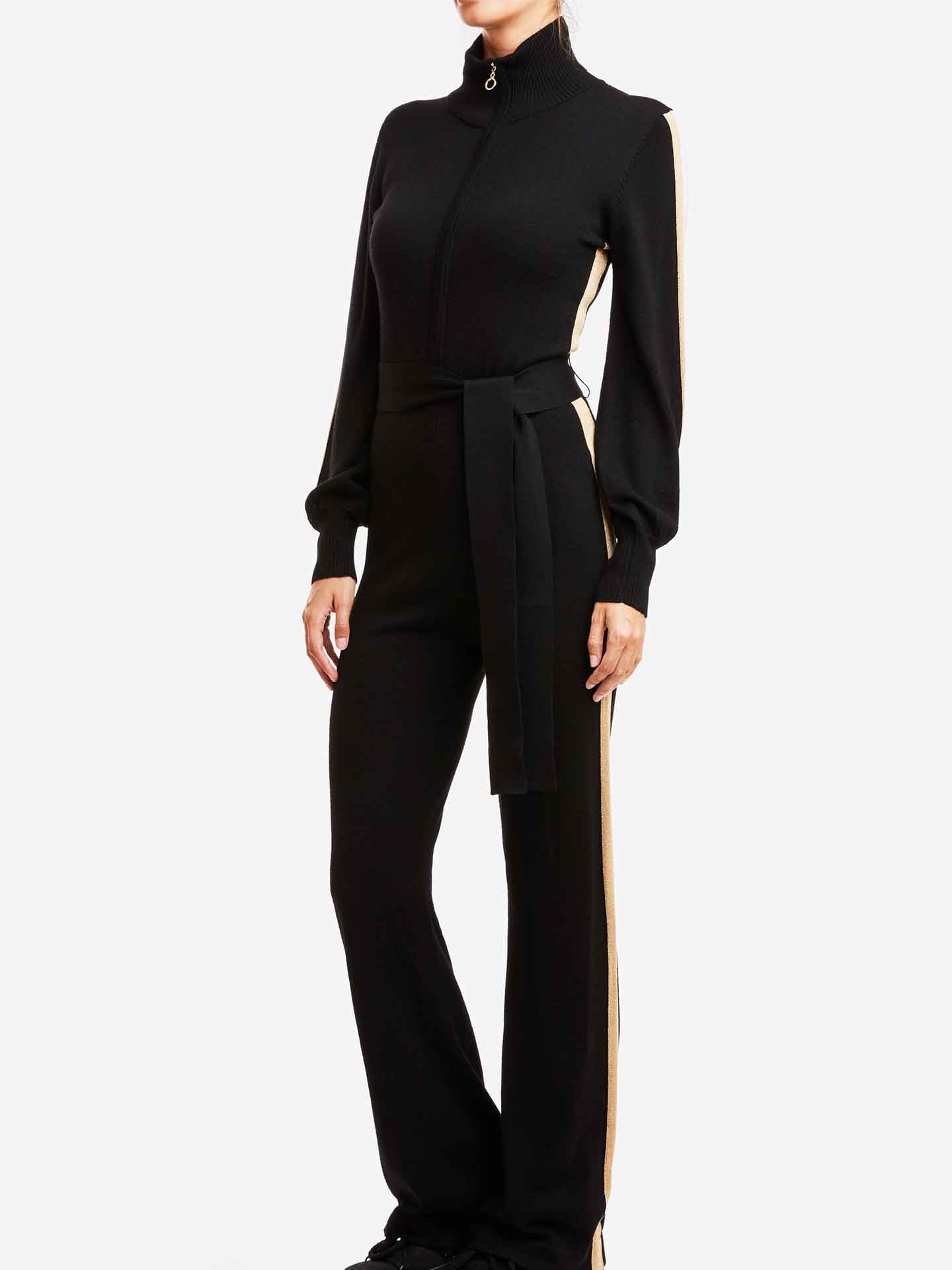 A person is modeling the stylish black Geilo Jumpsuit Women by We Norwegians, which boasts a high collar and long sleeves. This chic outfit features beige side stripes and a black belt tied at the waist, exuding a refined mountain resort vibe. The white backdrop accentuates the ensemble while keeping the model's head out of view.