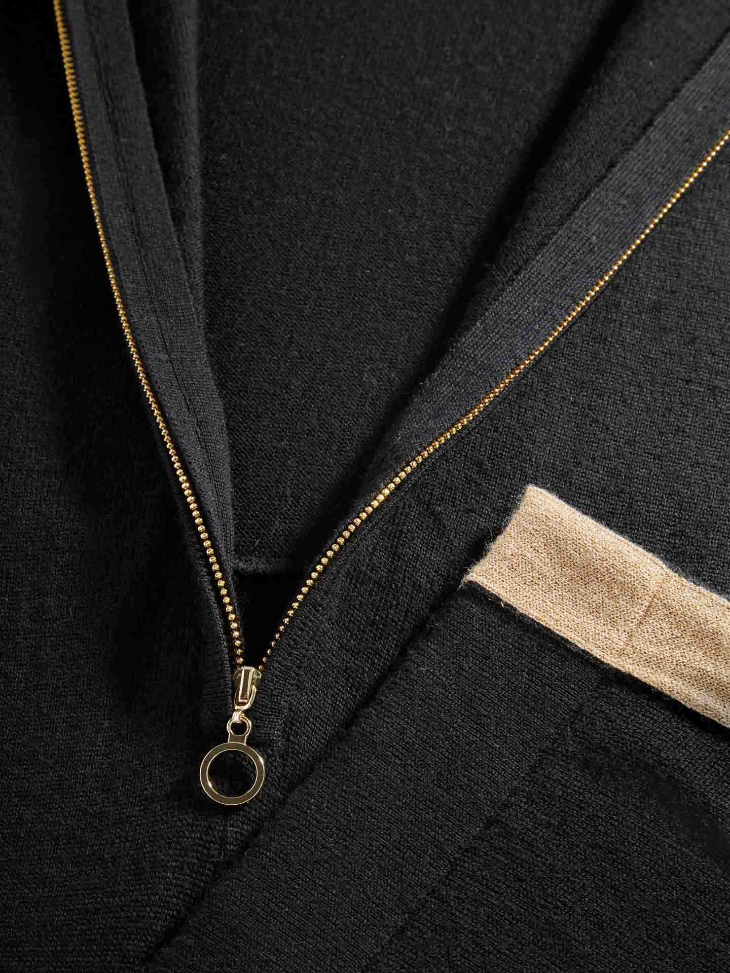 Close-up image of a Geilo Jumpsuit Women by We Norwegians, featuring a black zip-up design with a gold zipper. The zipper is partially undone, revealing the Merino Wool texture of the fabric. A beige-colored patch is visible near the zipper, possibly indicating a pocket or design element.