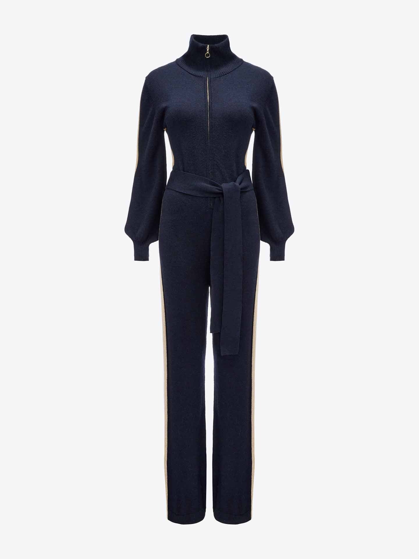 A full-body image showcases the elegant Geilo Jumpsuit Women by We Norwegians in navy blue with a high collar and zip-up front. Made from Merino Wool, it features long, puffed sleeves, wide cuffs, and a matching fabric belt tied around the waist. Beige stripes run down the sides of the legs, adding an exquisite touch perfect for a mountain resort.