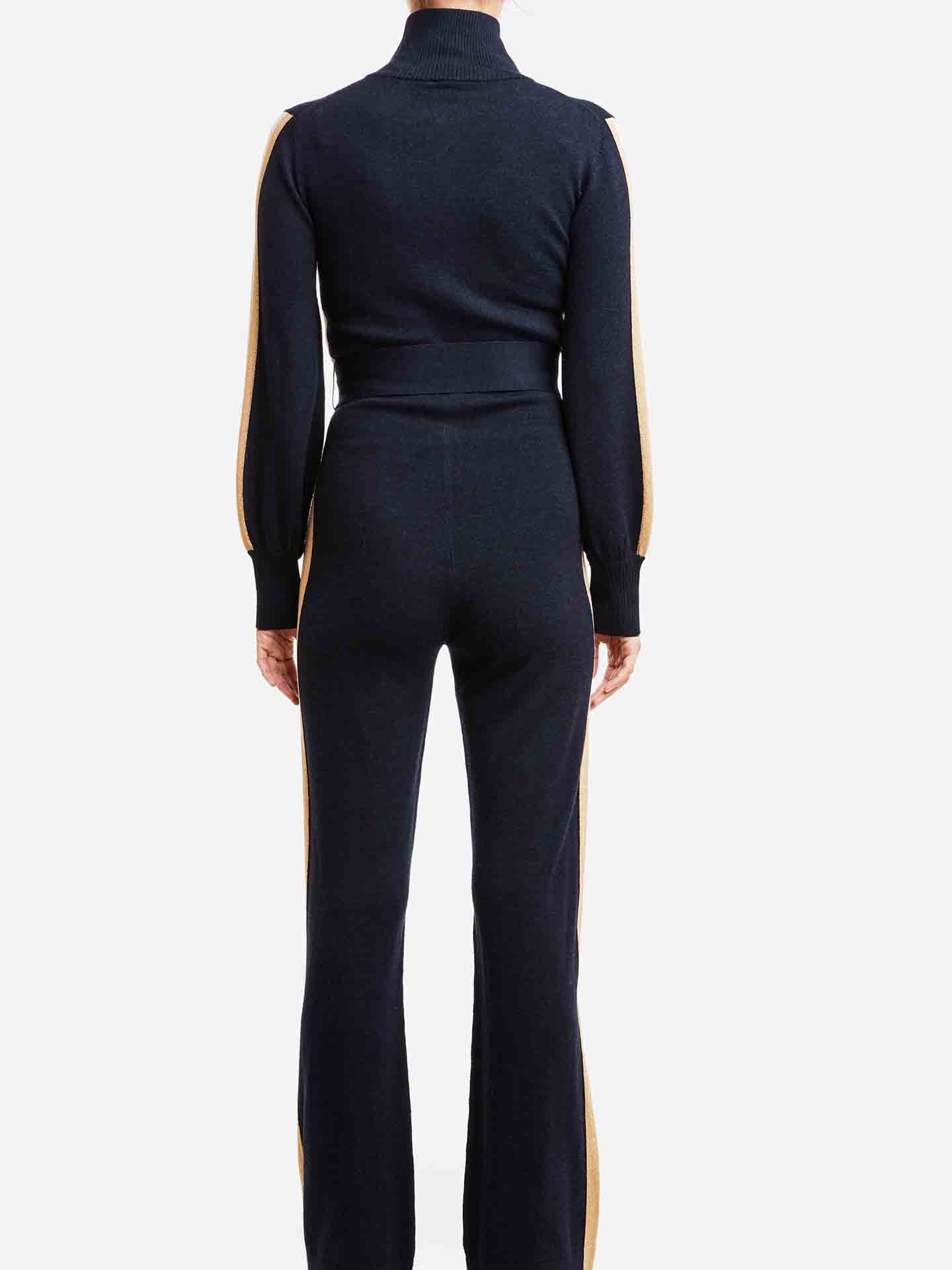 A person is standing with their back to the camera, wearing a chic Geilo Jumpsuit Women by We Norwegians, made from Merino wool. The outfit features a black sweater with a high collar and long sleeves, paired with matching pants adorned with yellow side stripes. The background is plain white.