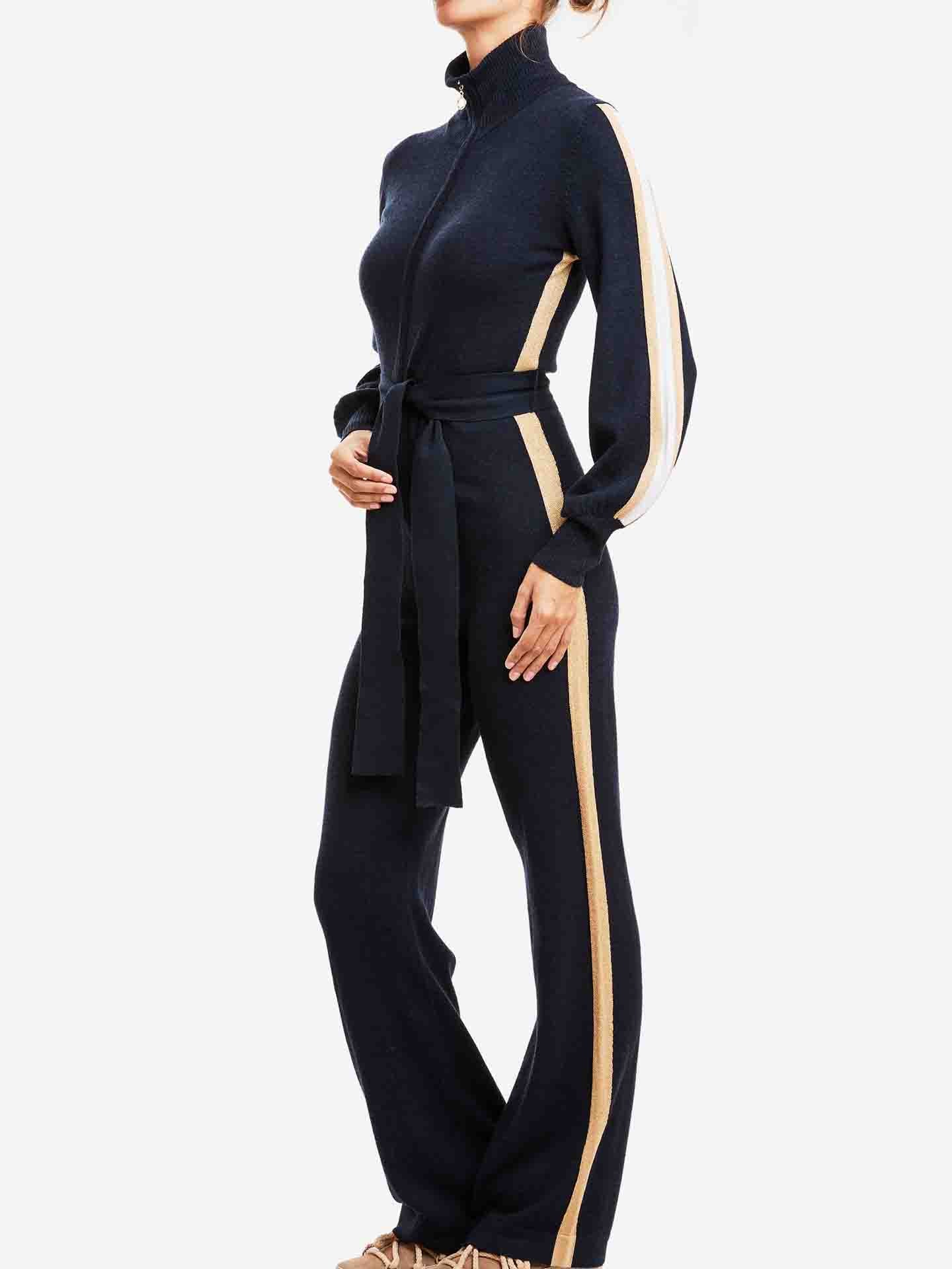 A woman is posing in a chic mountain resort, wearing a We Norwegians Geilo Jumpsuit made of Merino Wool. The full-body, dark-colored jumpsuit features vertical beige stripes on the sides of the arms and legs, paired with beige shoes and her hair tied back. It has a high collar and a fabric belt around the waist.