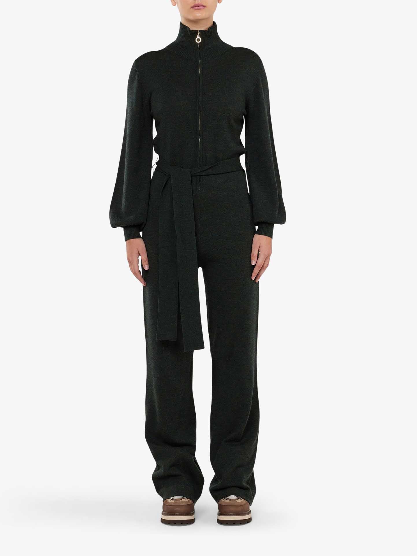 A person wearing the We Norwegians Geilo Jumpsuit Women in dark green, featuring a high collar, balloon sleeves, zipper front, and waist tie for the perfect chic mountain resort look, stands against a plain white background. The stylish ensemble is completed with brown shoes.