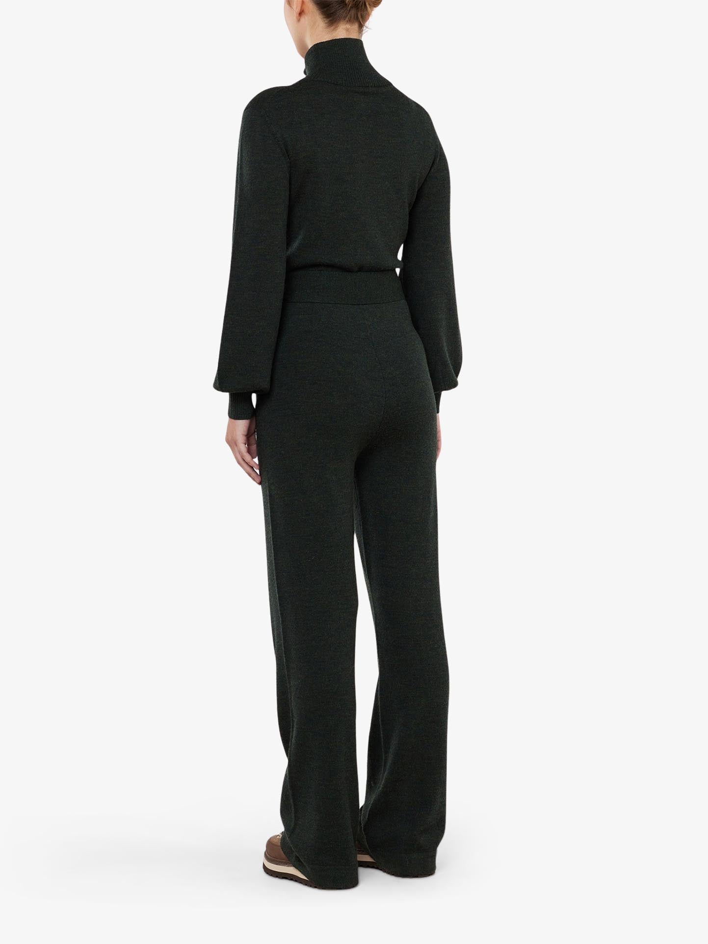 A person is shown from the back, wearing the Geilo Jumpsuit Women by We Norwegians, a chic dark green ensemble featuring a Fine Merino Wool turtleneck top and wide-leg pants. Their sophisticated mountain resort look is completed with brown shoes against a plain white background.