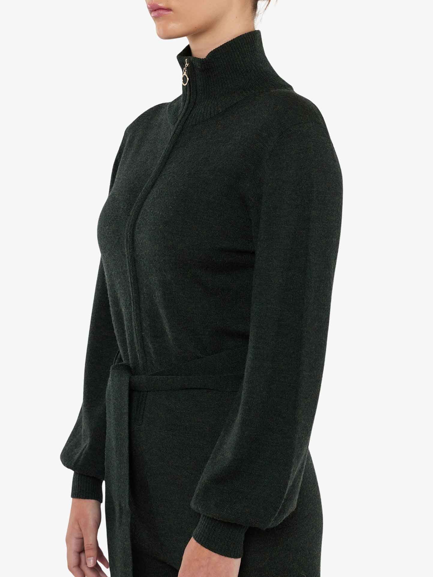 A person wearing the We Norwegians Geilo Jumpsuit for Women, featuring a dark green shade, long sleeves, high collar, zip front, and waist tie. The individual exudes chic mountain resort fashion while being shown from the side with their upper body and arms visible against a plain white background.