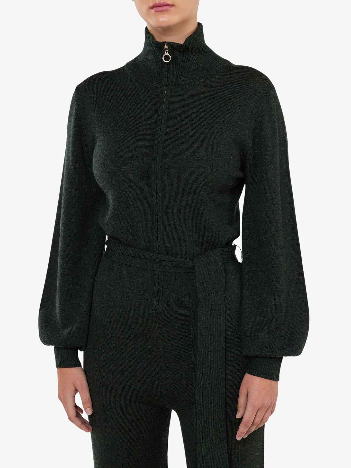 A person is wearing the We Norwegians Geilo Jumpsuit Women in dark green, featuring a high collar and front zipper. The jumpsuit includes a waist tie and slightly puffed sleeves, crafted from cozy Merino Wool. The background is plain white, and the person's face is not visible.