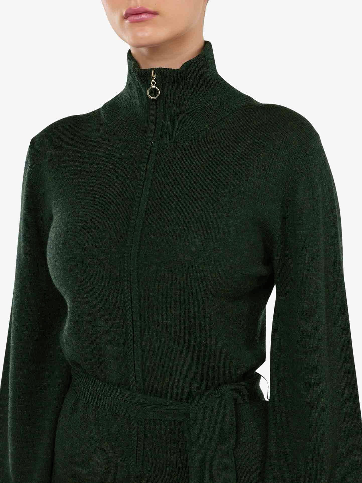 A person wearing the Geilo Jumpsuit by We Norwegians, featuring a dark green, zip-up, high-neck design with a waist tie that embodies chic mountain resort fashion. The cropped image highlights the upper torso and part of the face, focusing on the luxurious Merino Wool details against a plain white background.