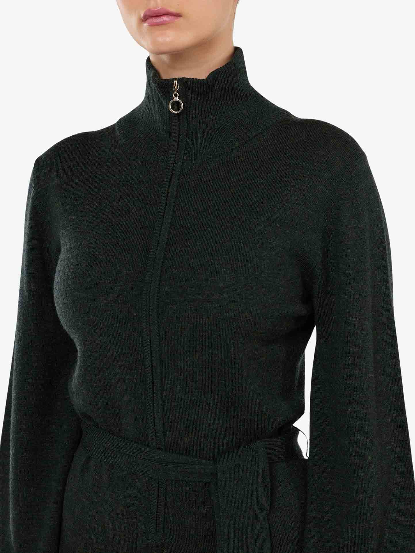 A person wearing a dark green Geilo Jumpsuit Women from We Norwegians stands against a plain white background. The full-zip jumpsuit features a high collar and matching belt around the waist, exuding the sophisticated charm of a chic mountain resort. The image captures only the upper half of the person's body.