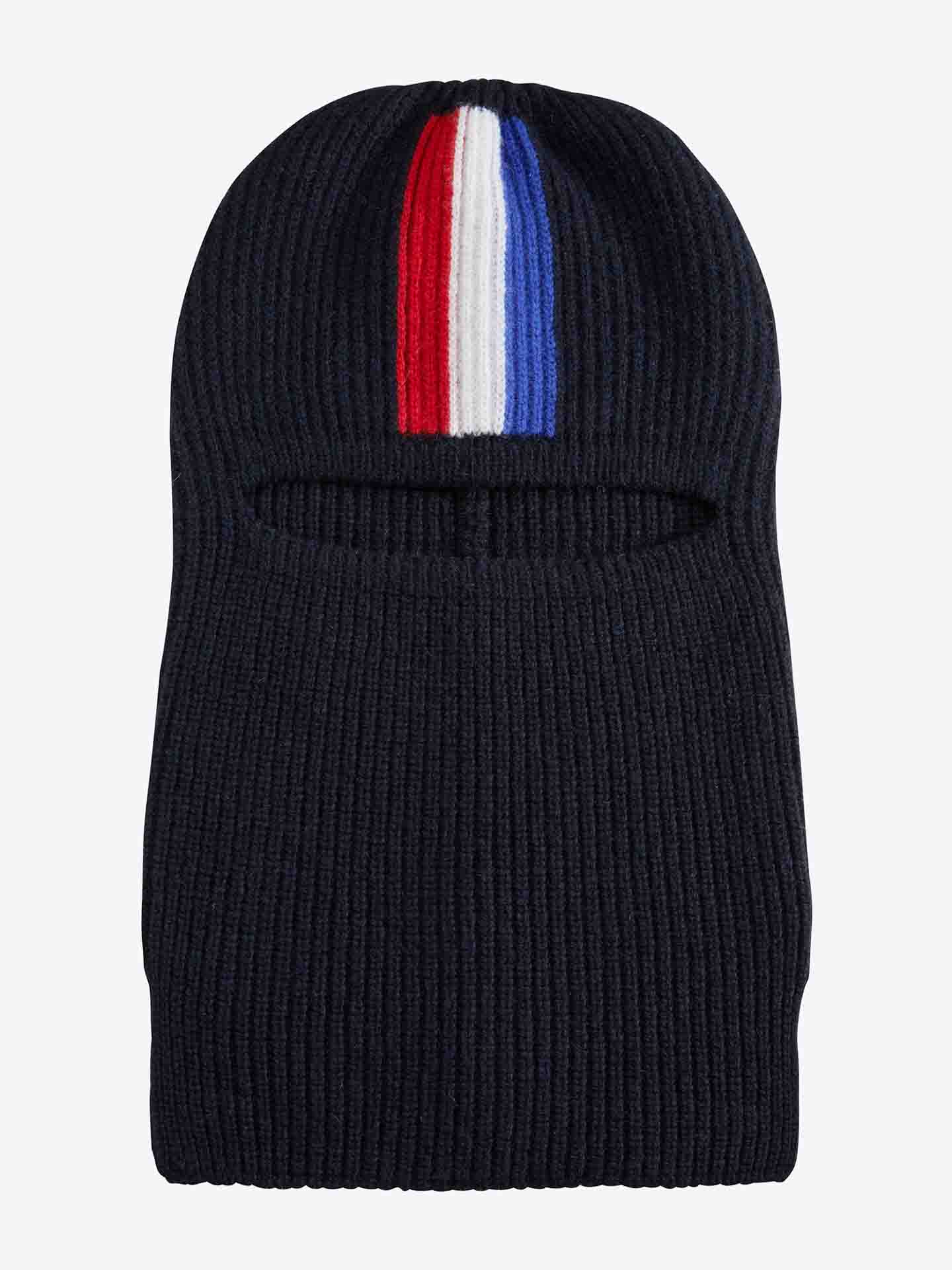 The We Norwegians Apres Ski Balaclava is a ribbed knit design crafted from dark merino wool, featuring an eye slit. The top center is adorned with a vertical stripe pattern in red, white, and blue, evoking the style of 70s ski fashion.