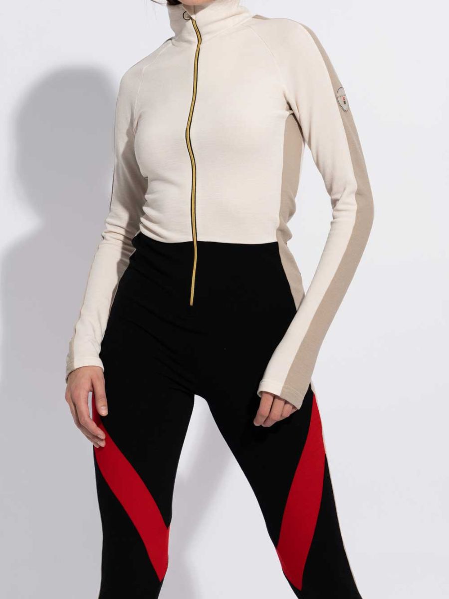 Dressed in the INUIKII + WN Jumpsuit for women by We Norwegians, paired with black leggings featuring red diagonal accents, she epitomizes après-ski elegance. Set against a plain white background, her sophisticated ski fashionista style is unmistakable.