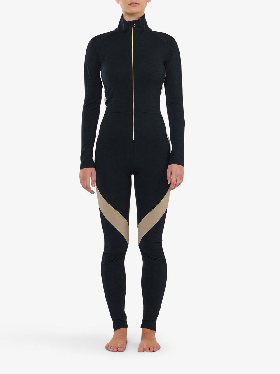 A person dressed in the stylish INUIKII + WN Jumpsuit Women by We Norwegians, featuring a sleek black design with a front zipper and beige diagonal stripes on the legs, stands barefoot against a plain white background, capturing the essence of ski fashionistas.