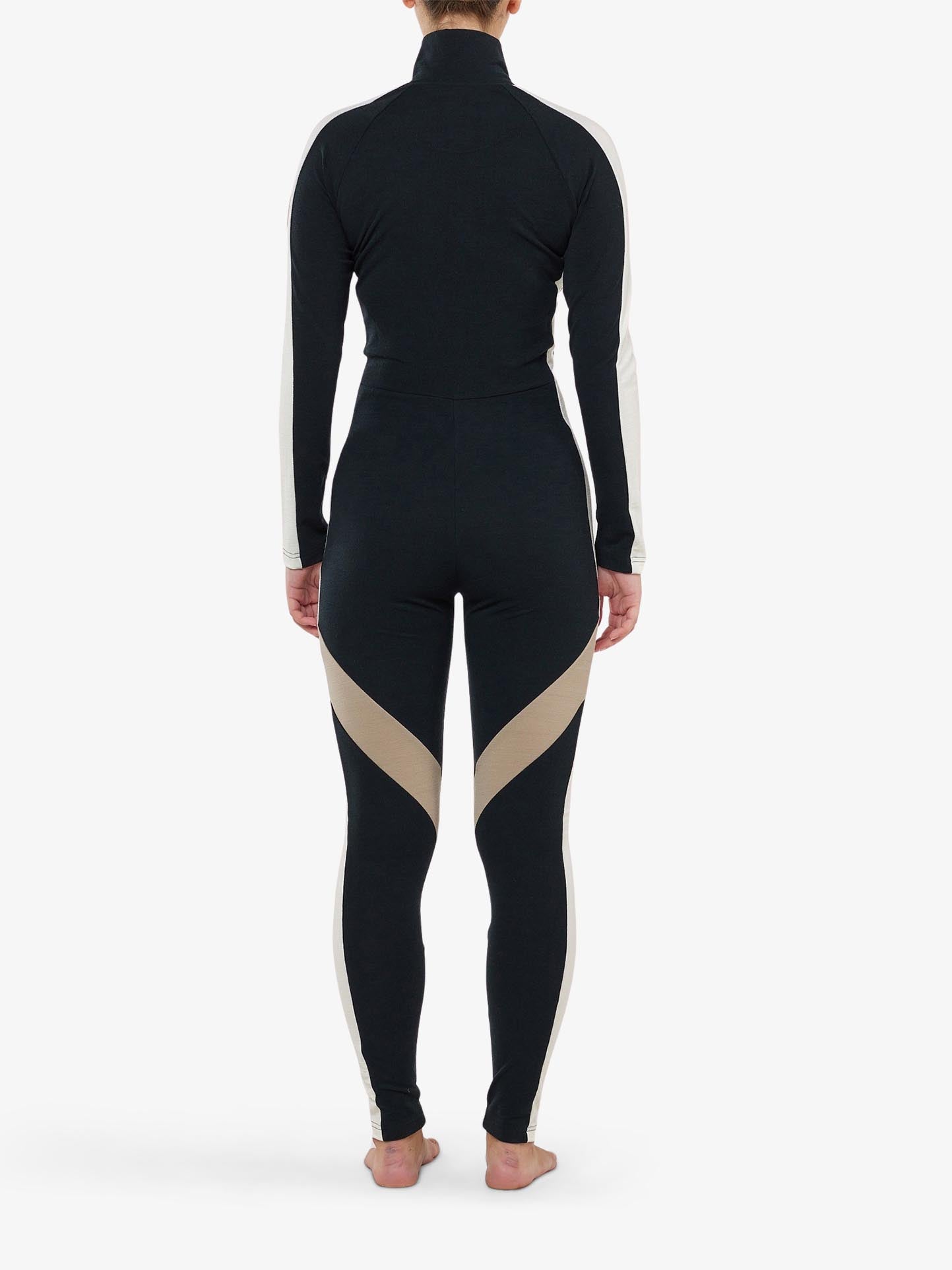 A person stands barefoot on a white background, presenting the INUIKII + WN Jumpsuit Women by We Norwegians in sleek black and beige. The jumpsuit features long sleeves and a high collar, with a photo capturing its elegant design from the back, epitomizing chic ski fashionista style.