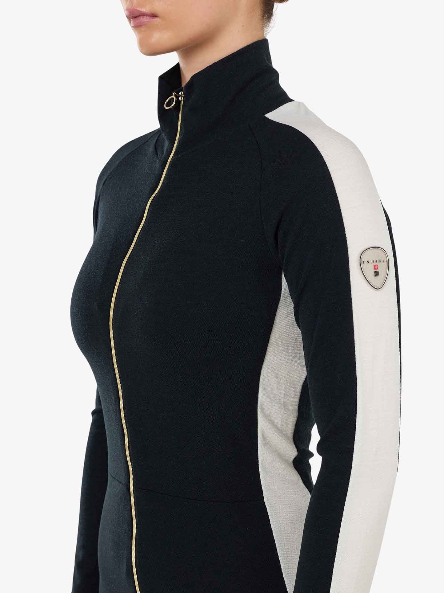 A person dressed in the INUIKII + WN Jumpsuit Women by We Norwegians, showcasing après ski style with a black zip-up top and distinct white sleeves. The high collar and gold zipper highlight its appeal to ski fashion aficionados, while a circular patch adorns the right sleeve. Only the upper torso and a profile view of the face are visible.