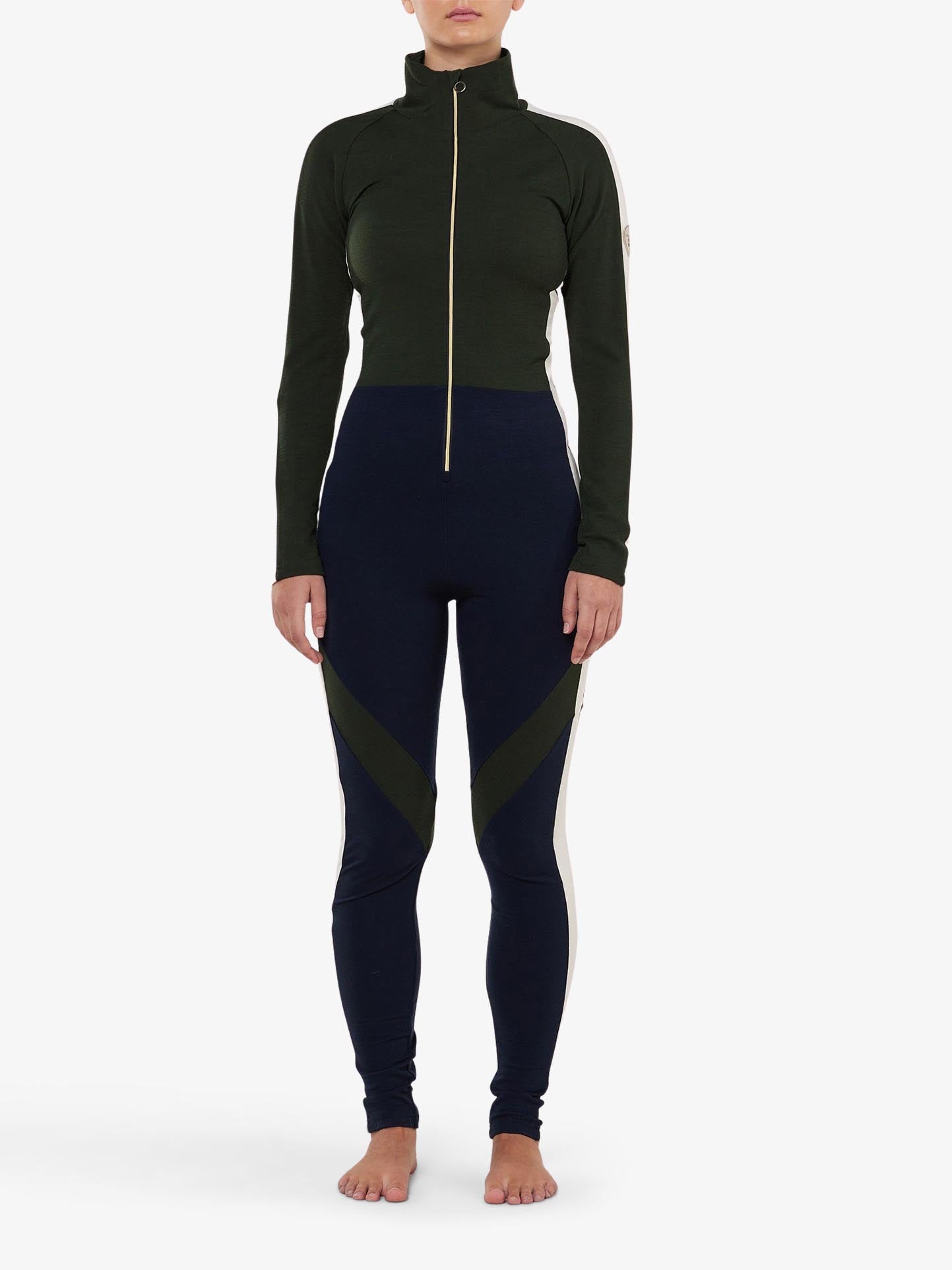 A person is wearing the We Norwegians INUIKII + WN Jumpsuit Women, a fitted athletic outfit with a high neck and long sleeves, featuring dark green and black colors with a front zipper. This ensemble exudes the flair of ski fashionistas and includes white stripes down the sleeves along with geometric patterns on the leggings. The individual stands barefoot against a plain backdrop.