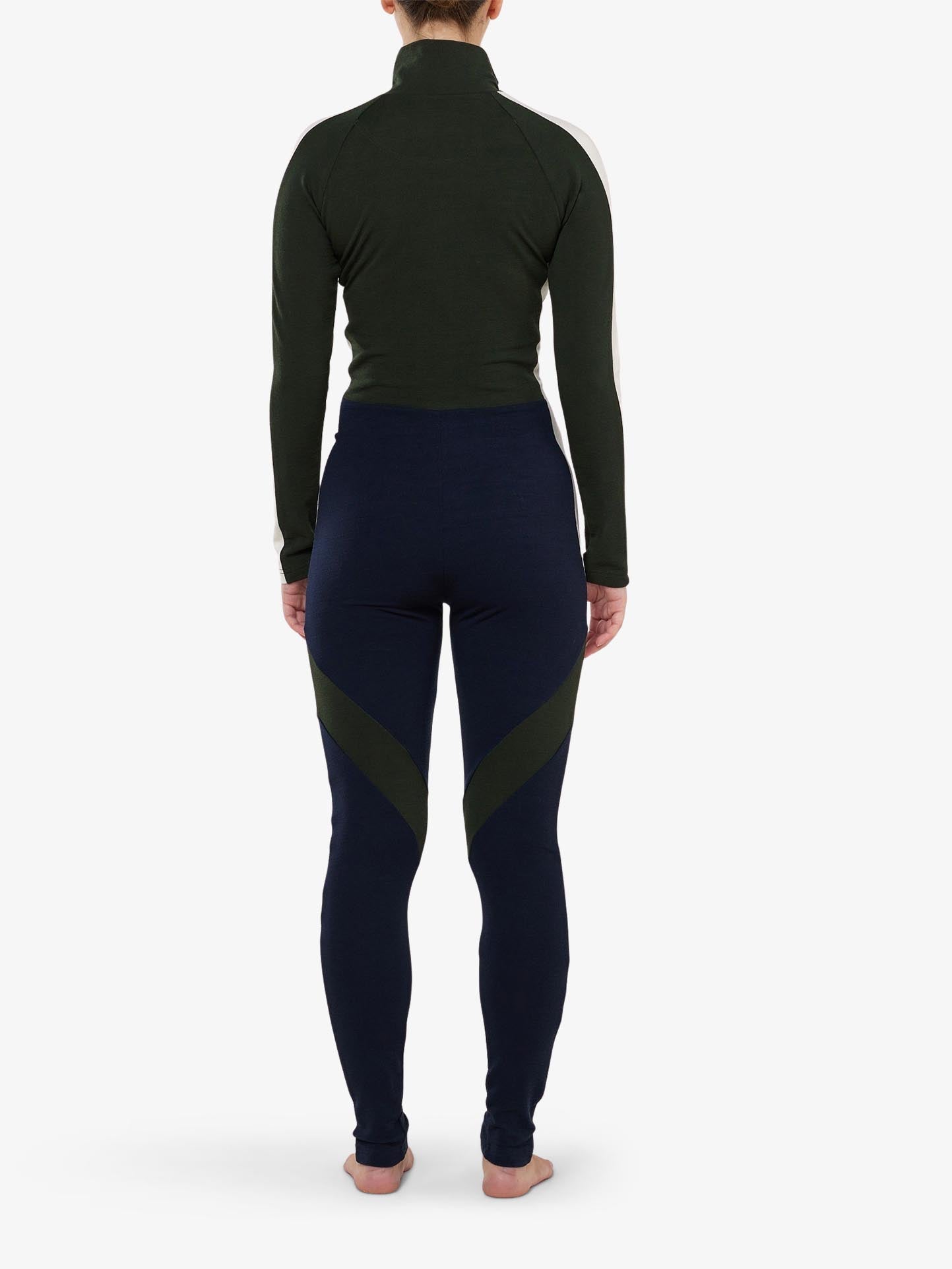 A person stands facing away, exuding ski fashionista vibes in the INUIKII + WN Jumpsuit by We Norwegians, showcasing a dark green high-neck top and navy leggings with green accents. They are barefoot on a white background, embracing an aprés ski elegance.