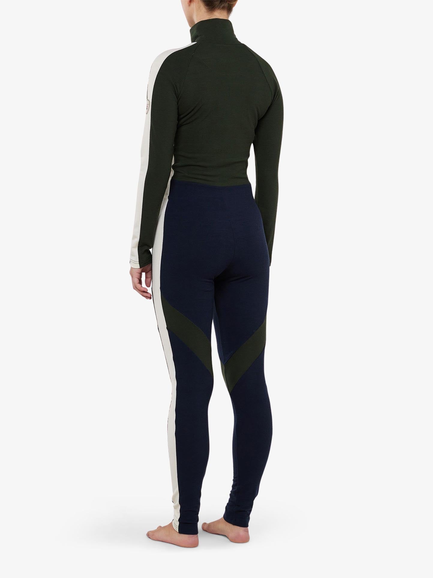 A person is barefoot, turned away, wearing the INUIKII + WN Jumpsuit Women by We Norwegians, emitting ski fashionista vibes. This form-fitting, long-sleeve jumpsuit features a color-blocked design with a dark green and cream top paired with navy leggings highlighted by green accents. The backdrop is plain white.