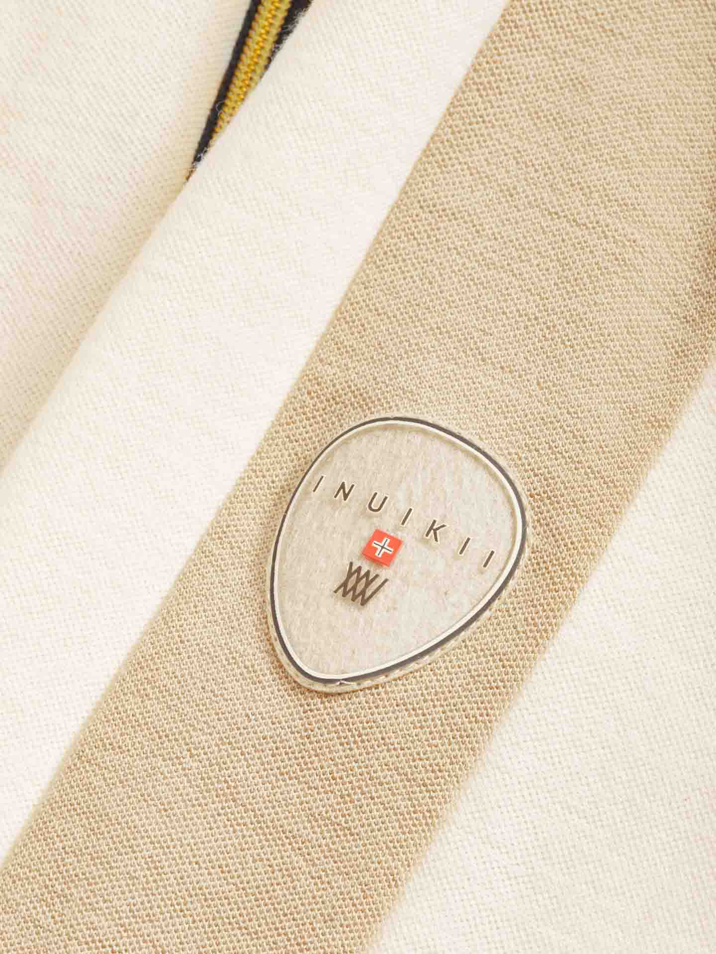 The INUIKII + WN Jumpsuit Women by We Norwegians showcases a close-up of its textured fabric in neutral tones, adorned with an oval-shaped INUIKII logo badge in silver and red—a subtle nod to ski fashionistas. The background presents a pattern of off-white and beige stripes, evoking the sophisticated allure of an après-ski setting.